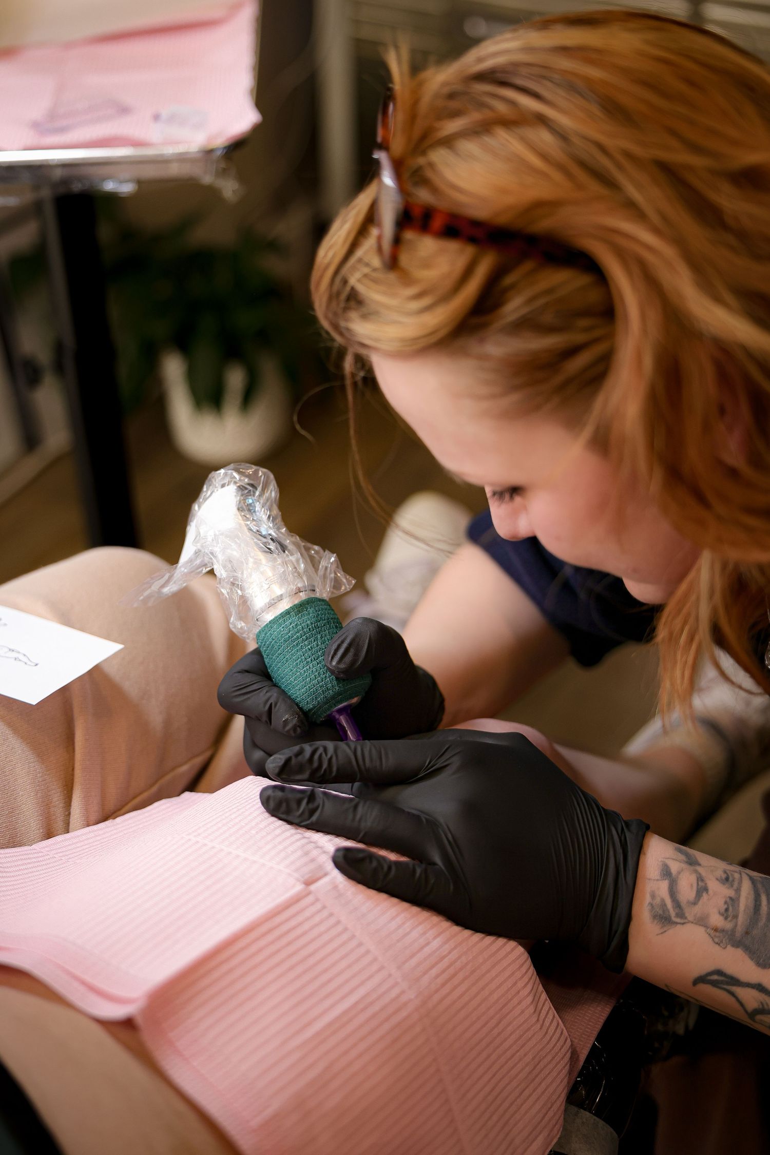 Red-haired tattoo artist wearing black gloves carefully applies ink using a tattoo machine in a professional studio setting.