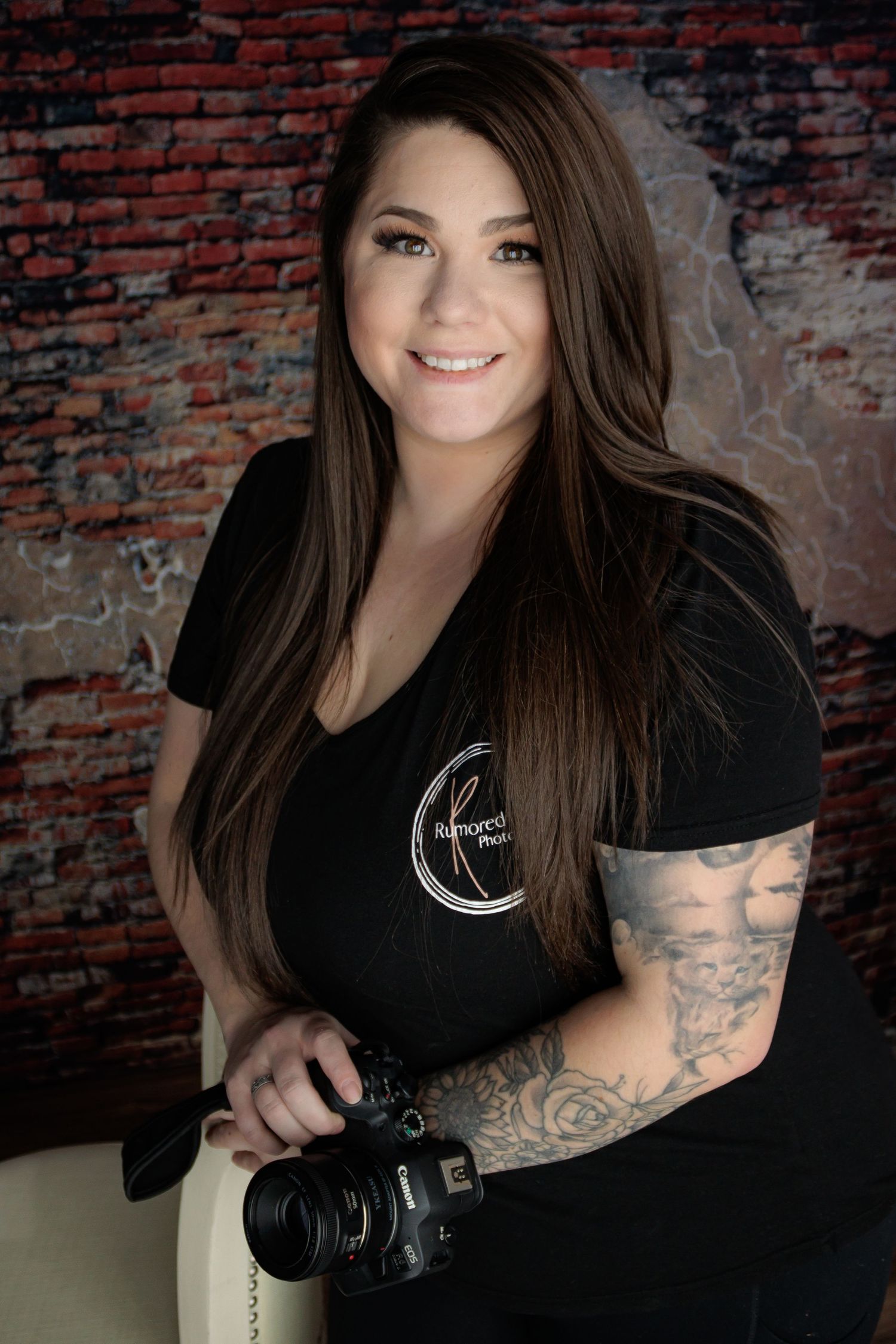 Professional photographer in black shirt with tattoos poses with camera against brick wall.
