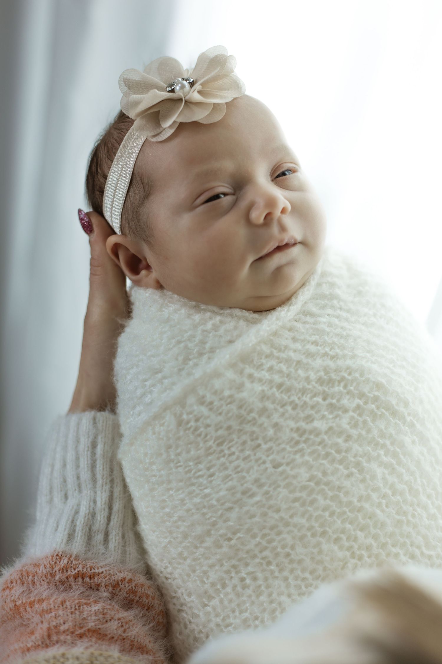 Baby wrapped in white knit blanket with bow headband in soft window light.