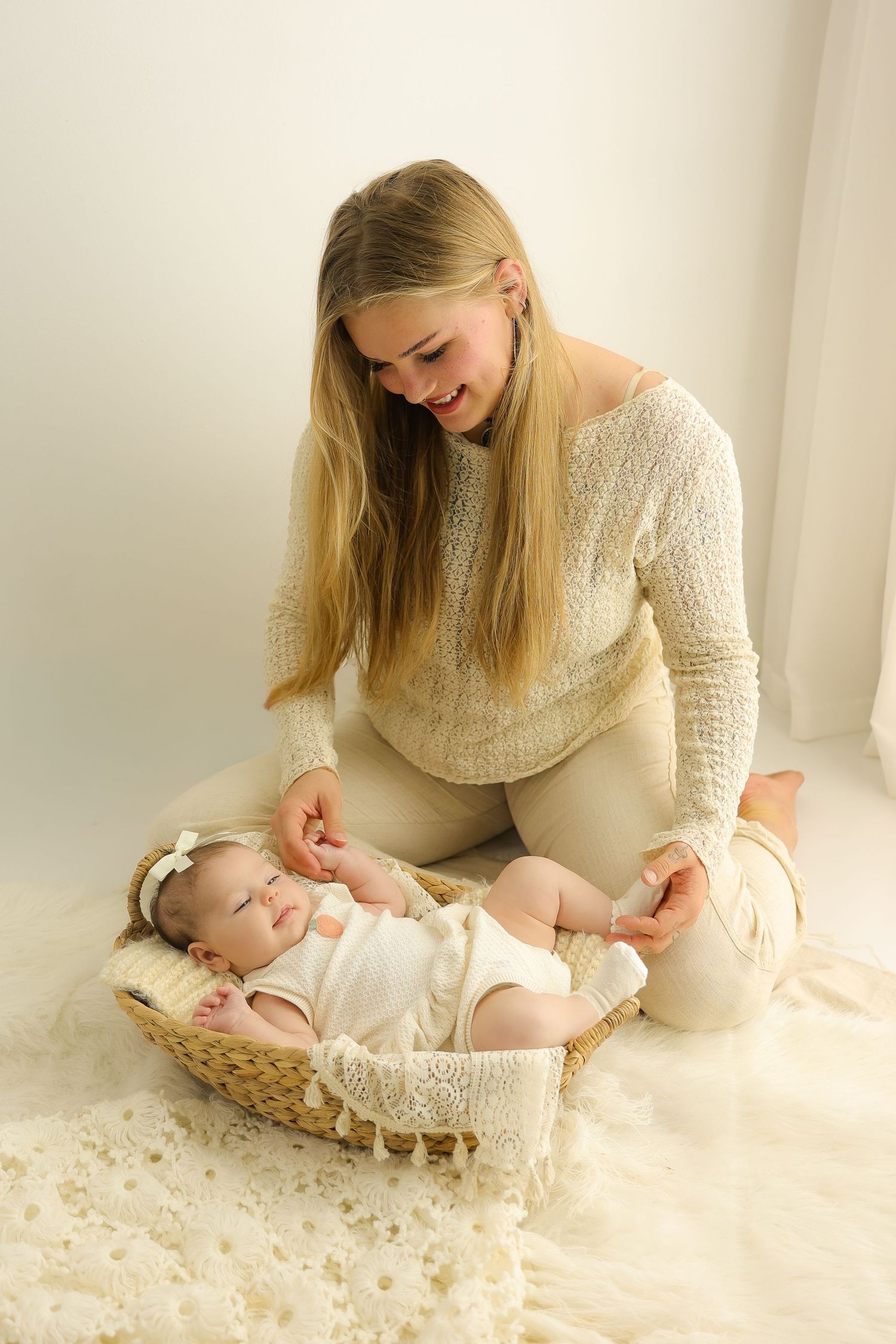 Smiling blonde mother kneeling on white fur rug, tenderly attending to newborn baby lying in wicker basket.