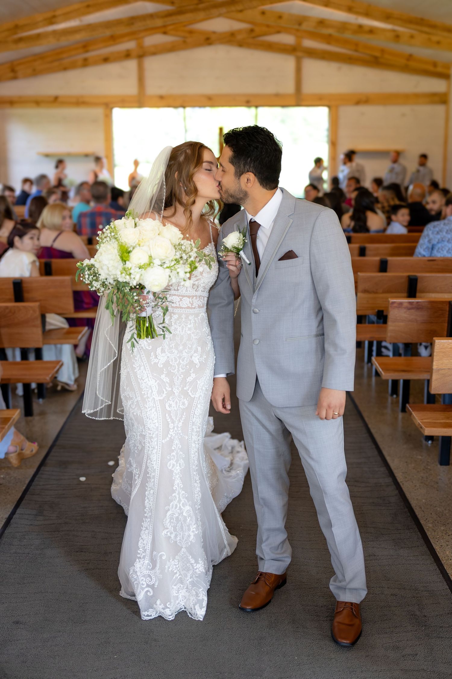 A bride in a lace gown and groom in a light gray suit share their first kiss as newlyweds in a rustic chapel.