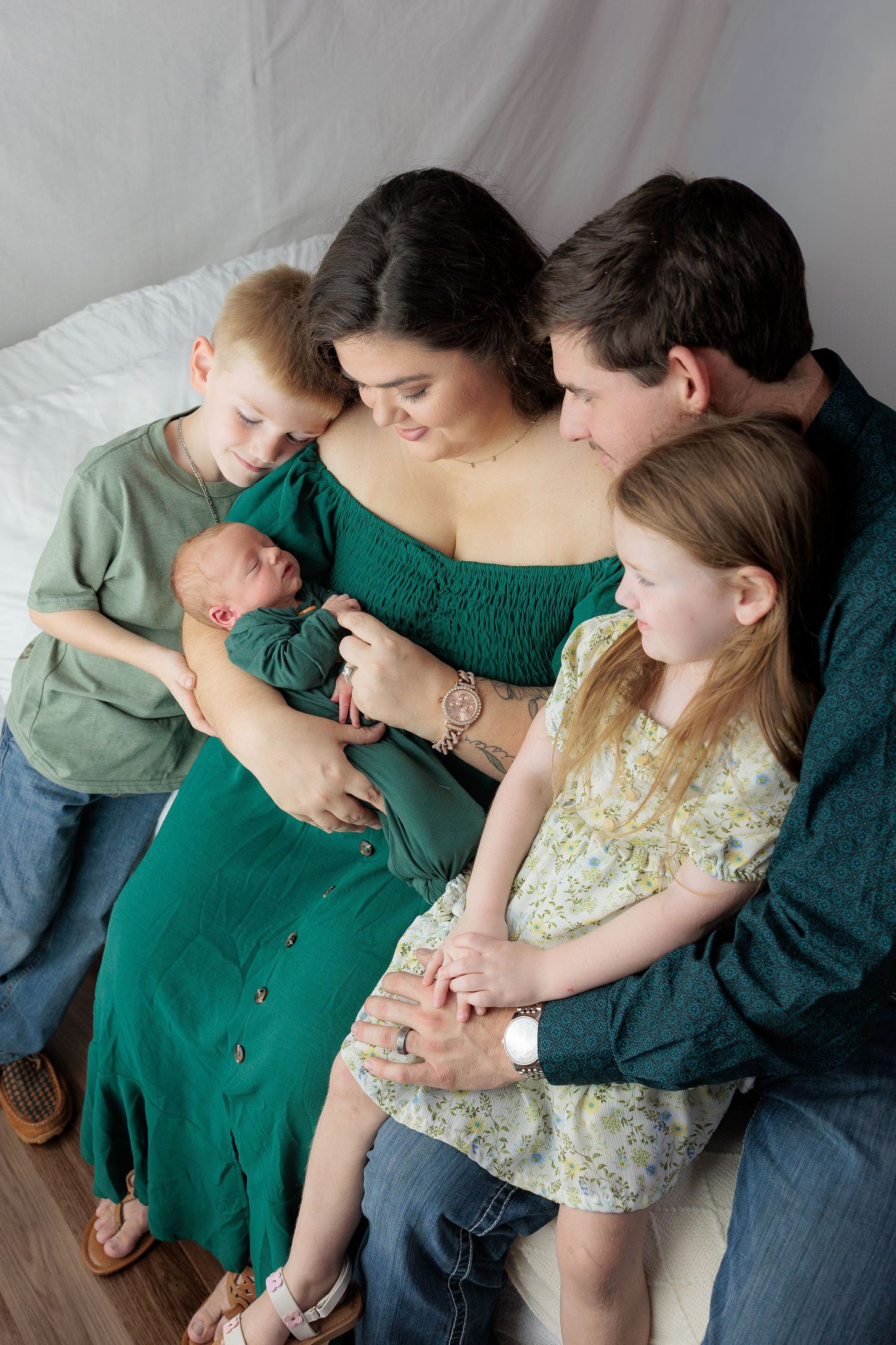 A family sits together on a couch, wearing green and floral outfits while sharing an intimate moment together.
