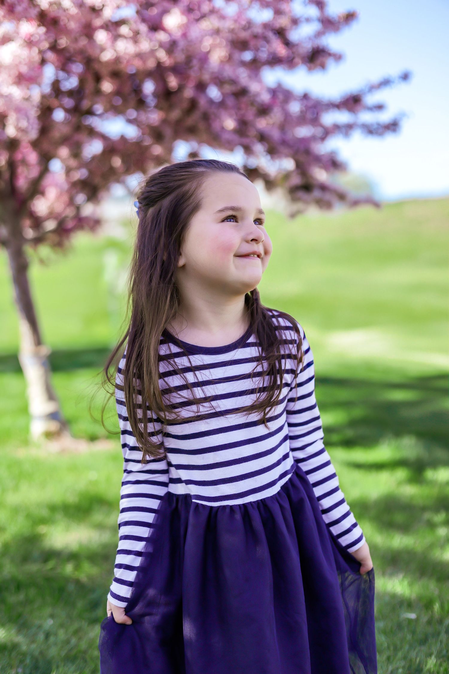 A child in a navy and white striped dress smiles while standing under blooming cherry blossom trees.
