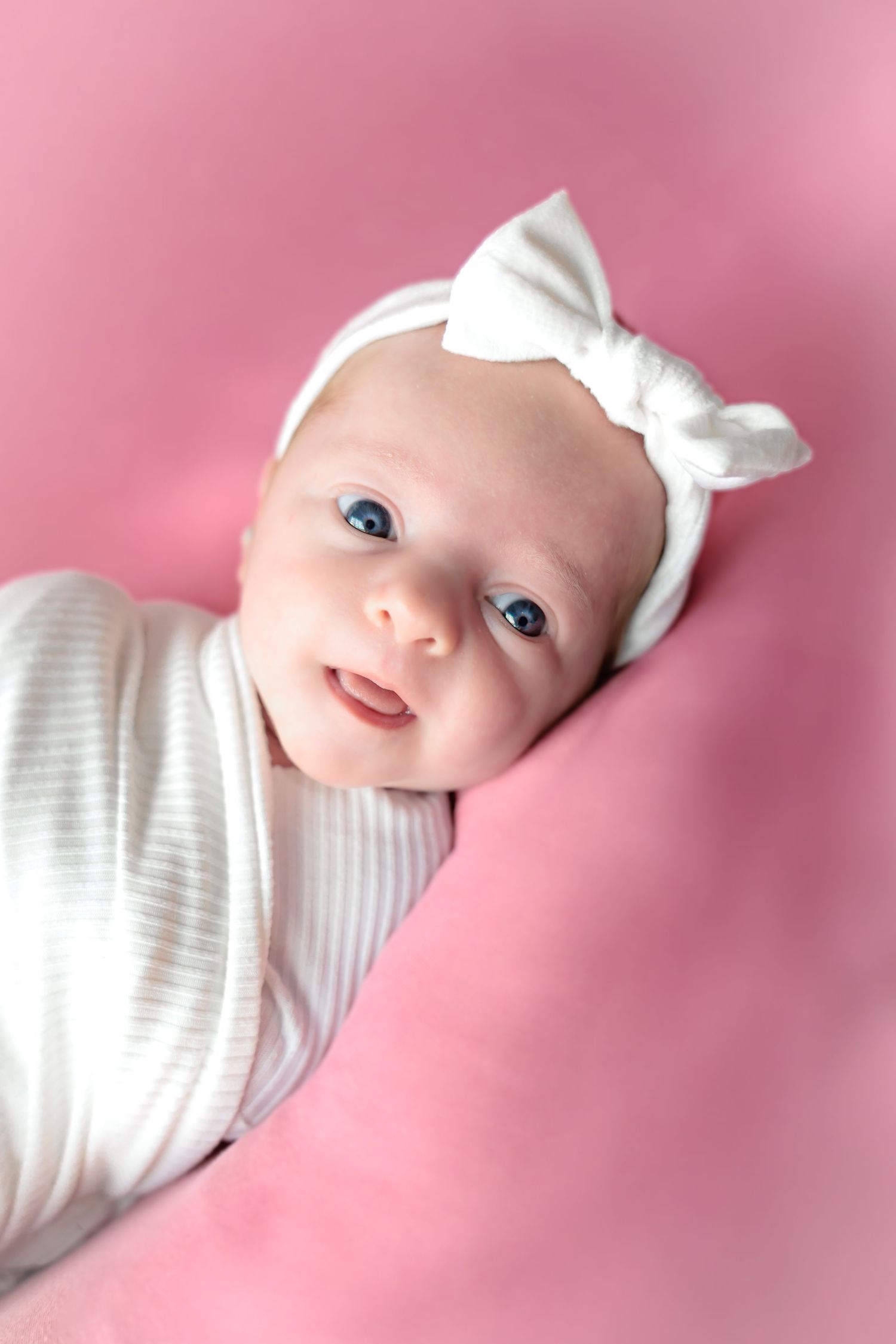 Sweet baby with a white bow lies on pink background looking at camera with bright blue eyes.
