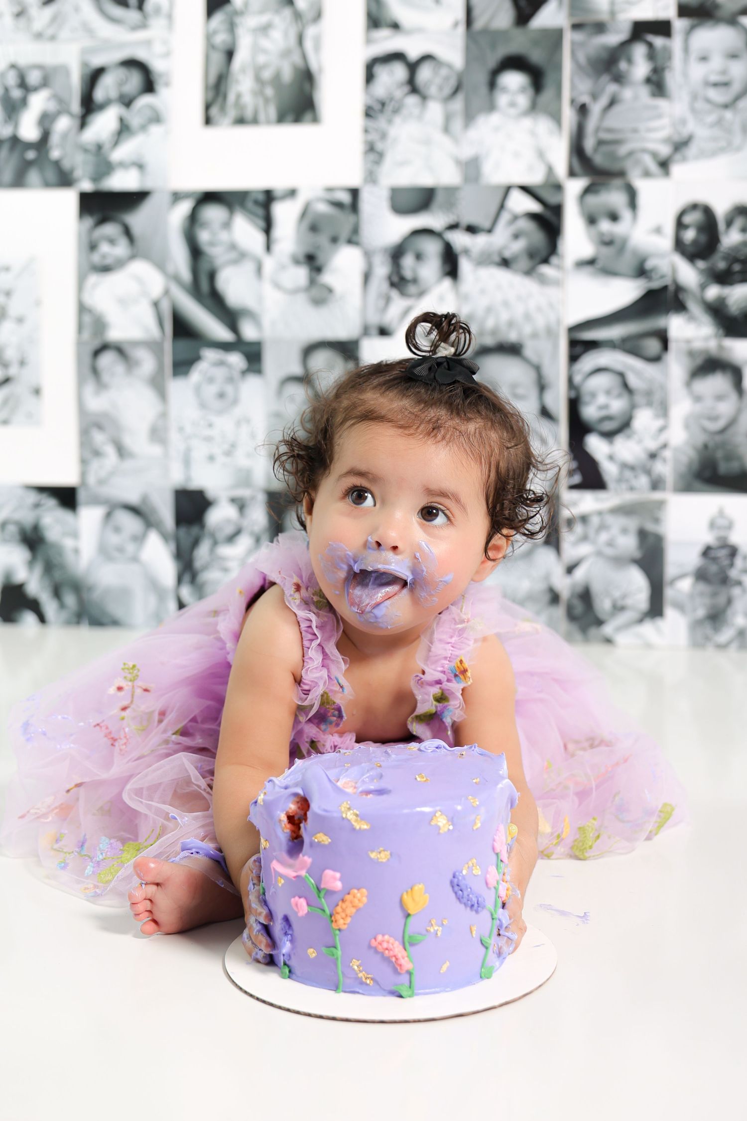 Baby in purple dress enjoying cake smash photoshoot with vintage photo wall background.