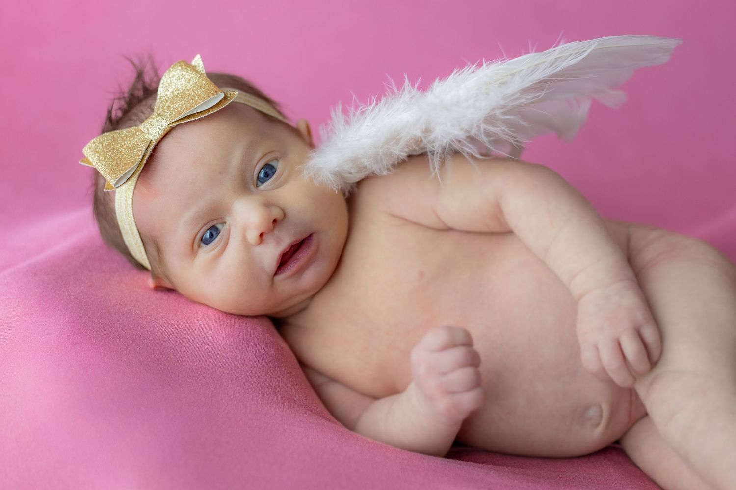 Baby wearing gold bow headband and angel wings on pink background.
