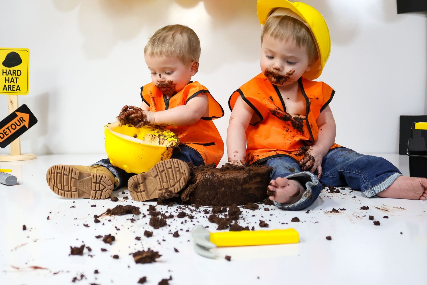 Baby in construction outfit playing with chocolate cake on white floor.