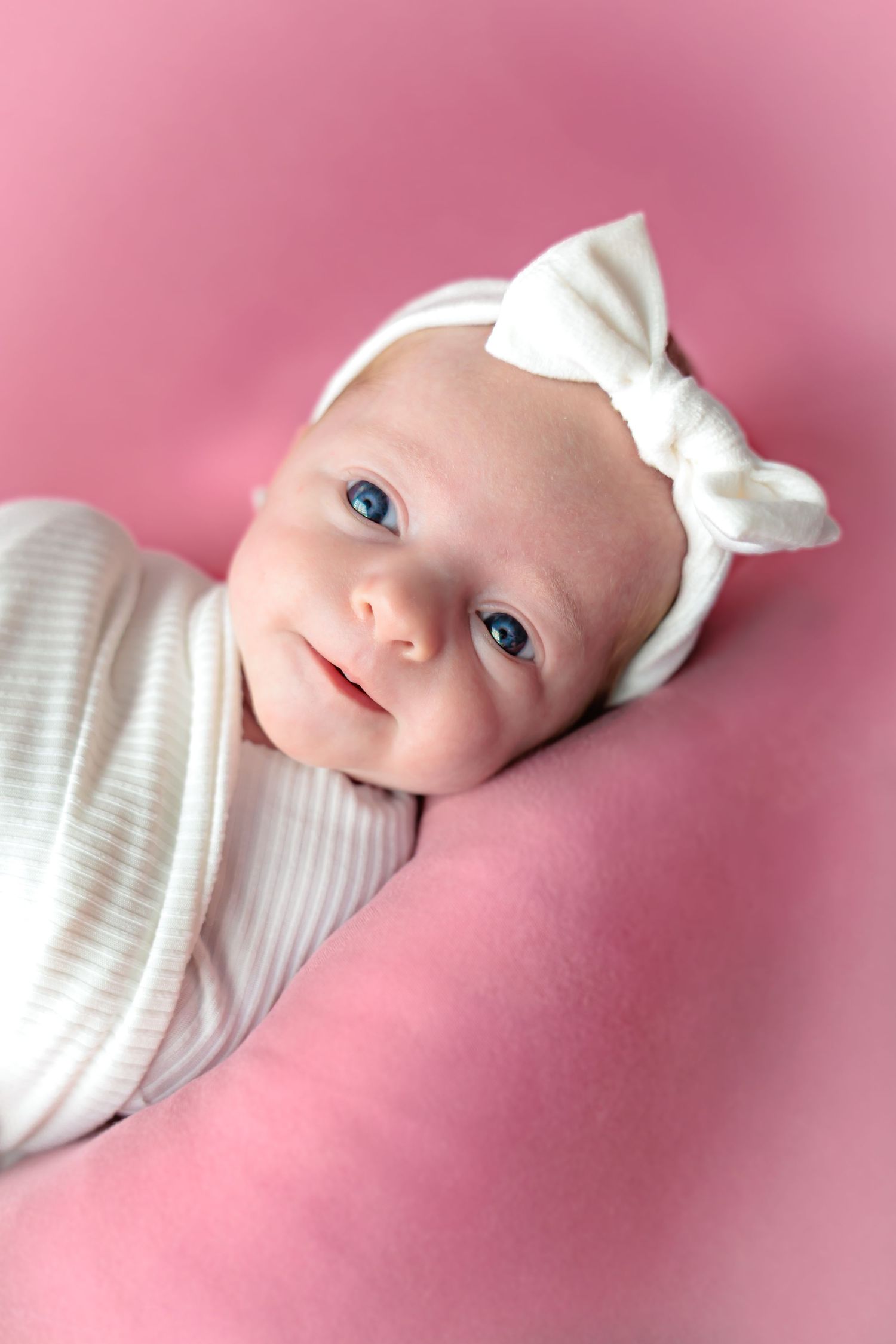 Baby wearing white bow headband on pink background with bright blue eyes.