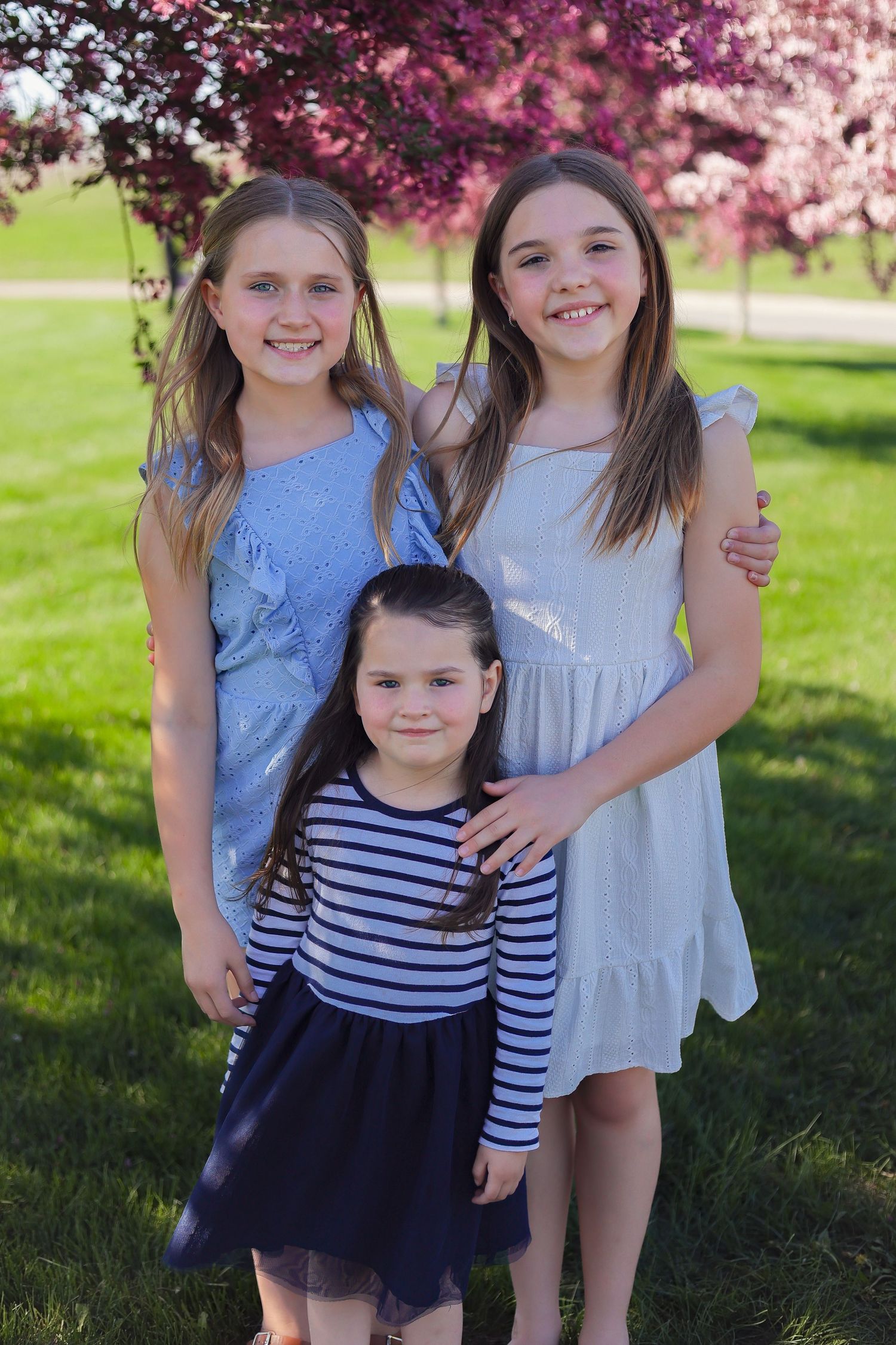 Three children pose together under blooming cherry trees in springtime.