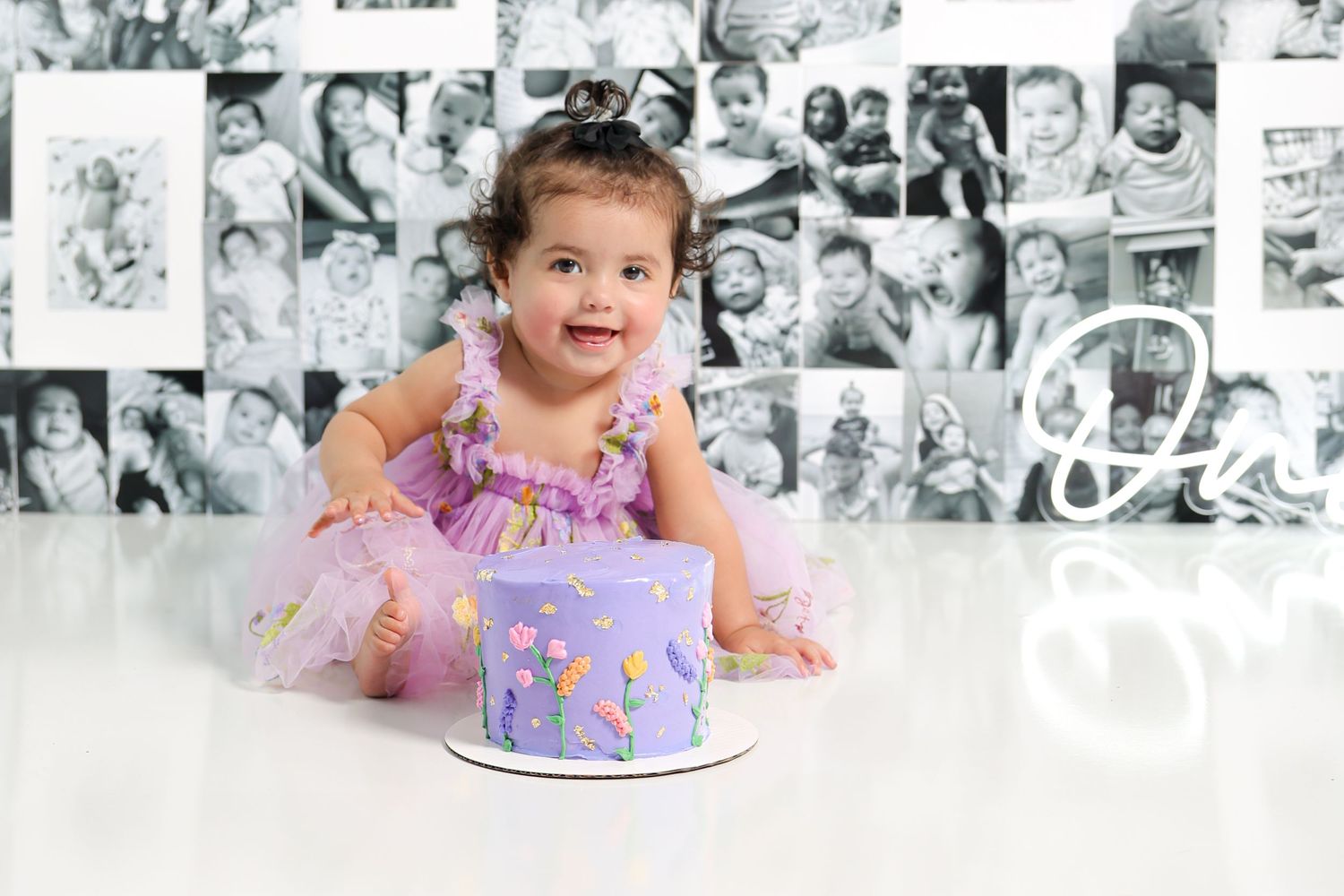 Baby in tulle dress reaches toward purple cake with black and white photo wall backdrop.