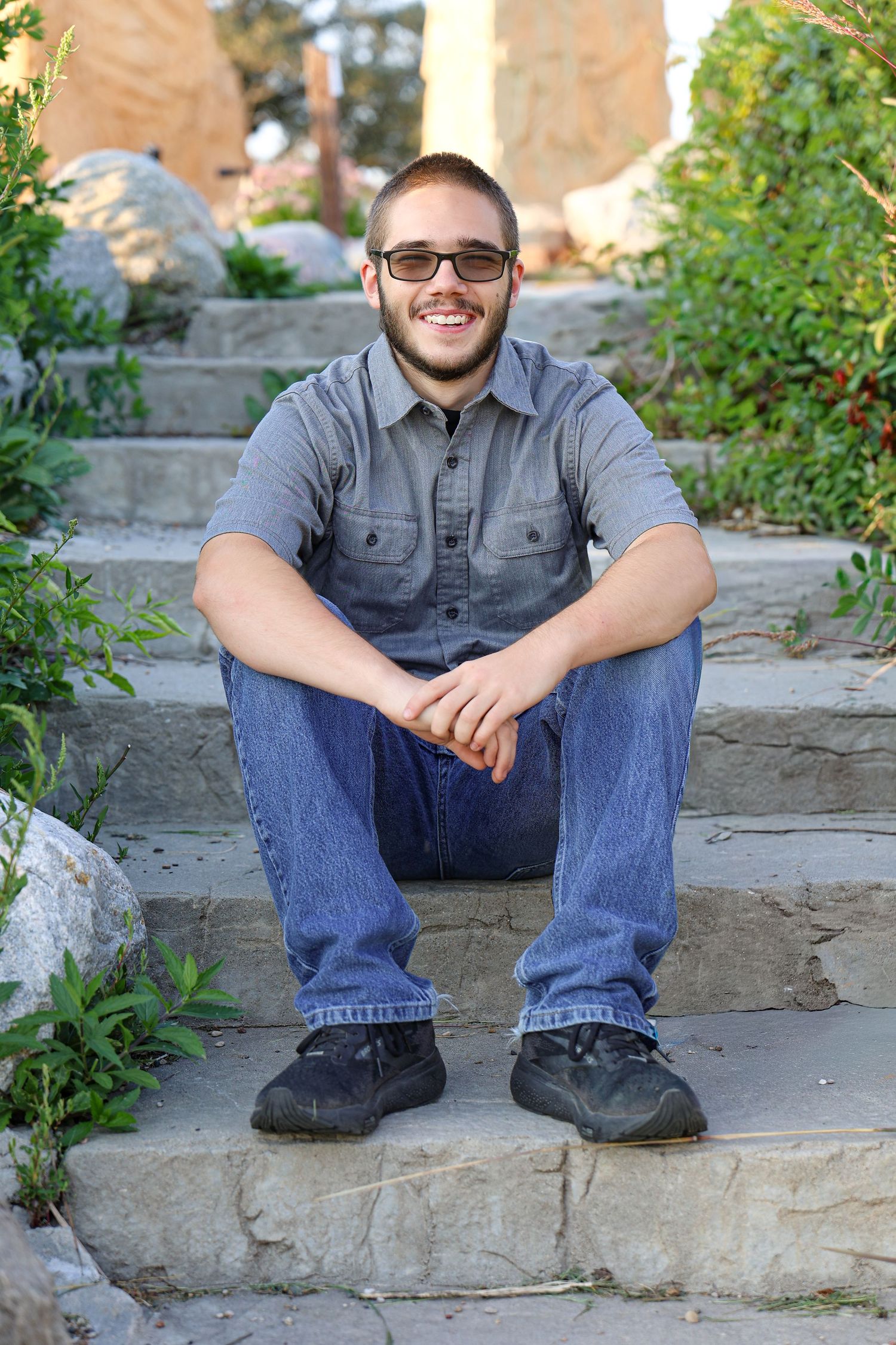 Person in casual attire sits on stone steps wearing a grey shirt, jeans and glasses in an outdoor setting.