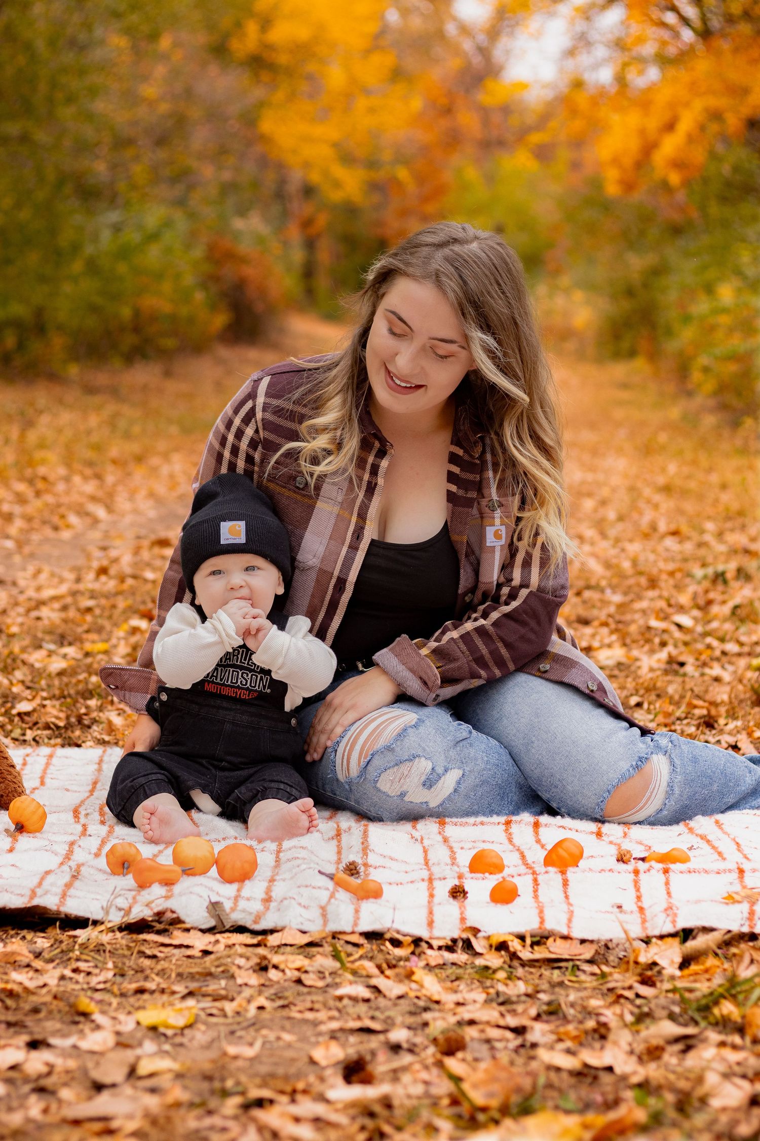 Mother and baby having autumn photoshoot on blanket with pumpkins and fall foliage.