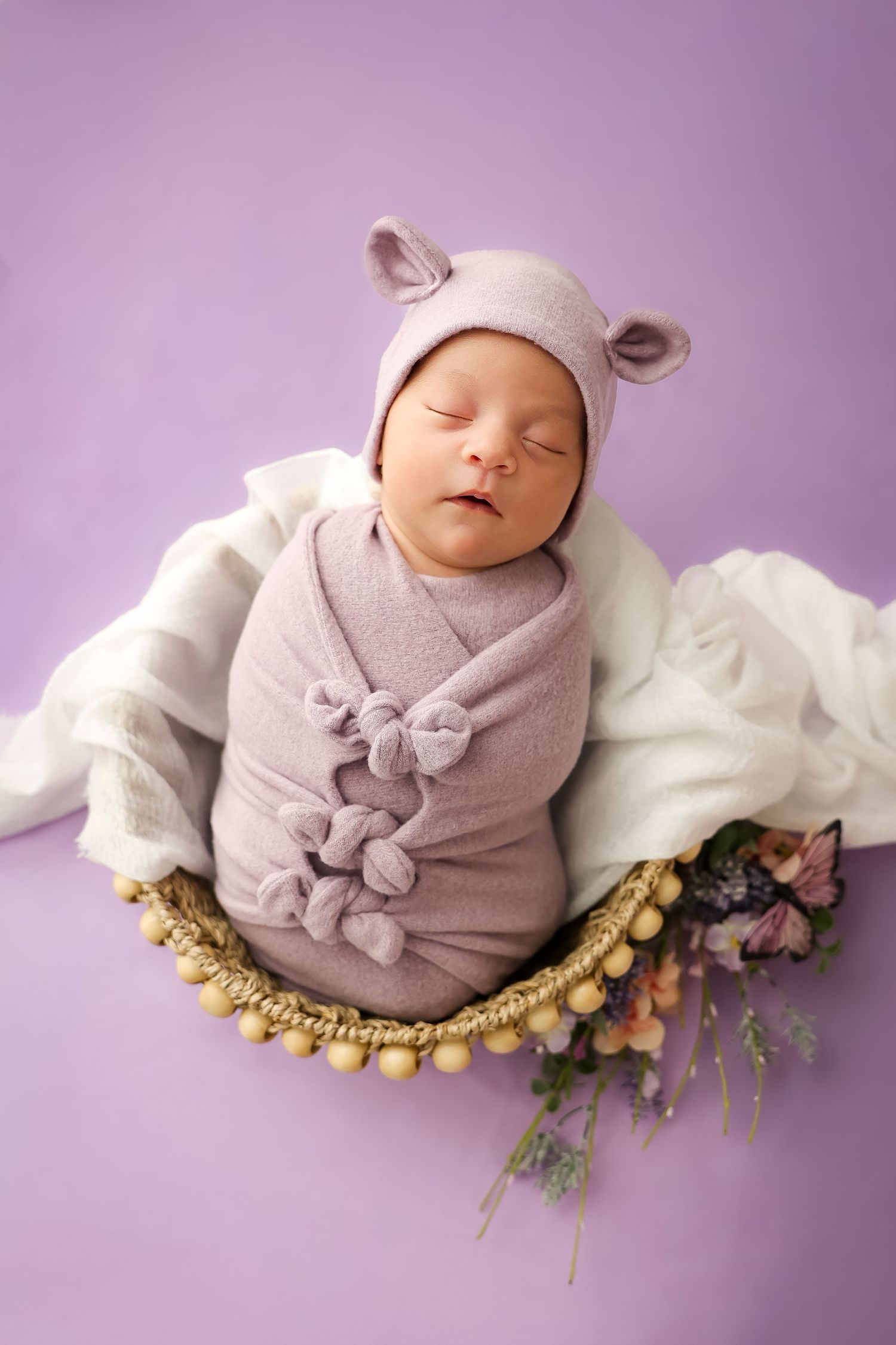 Newborn baby swaddled in lavender wrap wearing bear ear bonnet, sleeping in decorative basket with flowers on purple background.