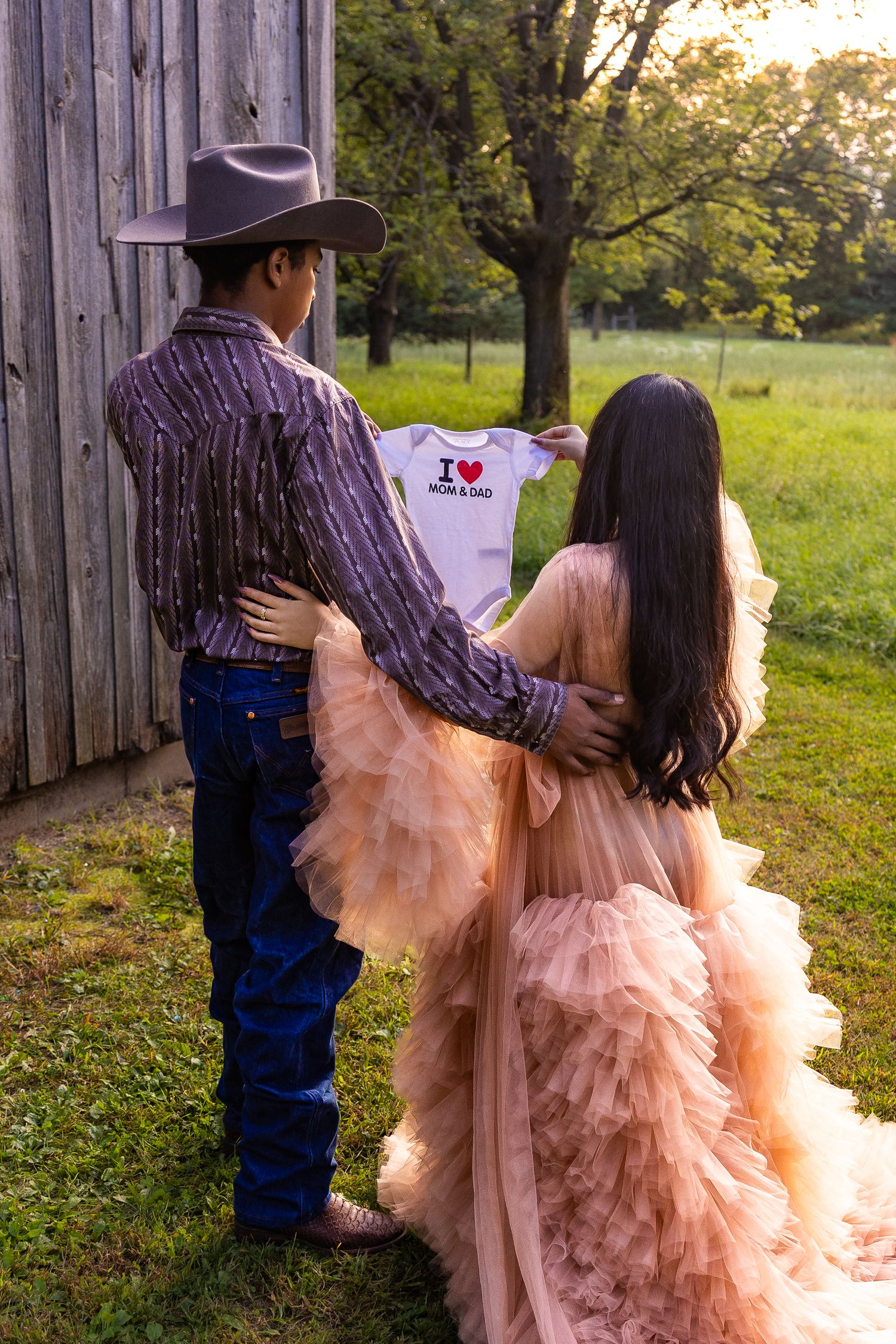A couple in western wear and pink tulle dress share intimate moments against a rustic barn wall at sunset.