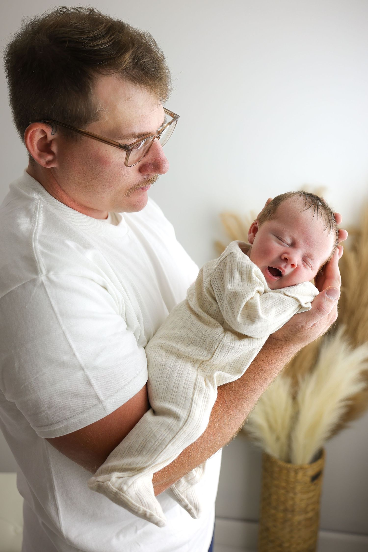 A parent in a white shirt holds and comforts a crying baby in a white outfit against a light background.