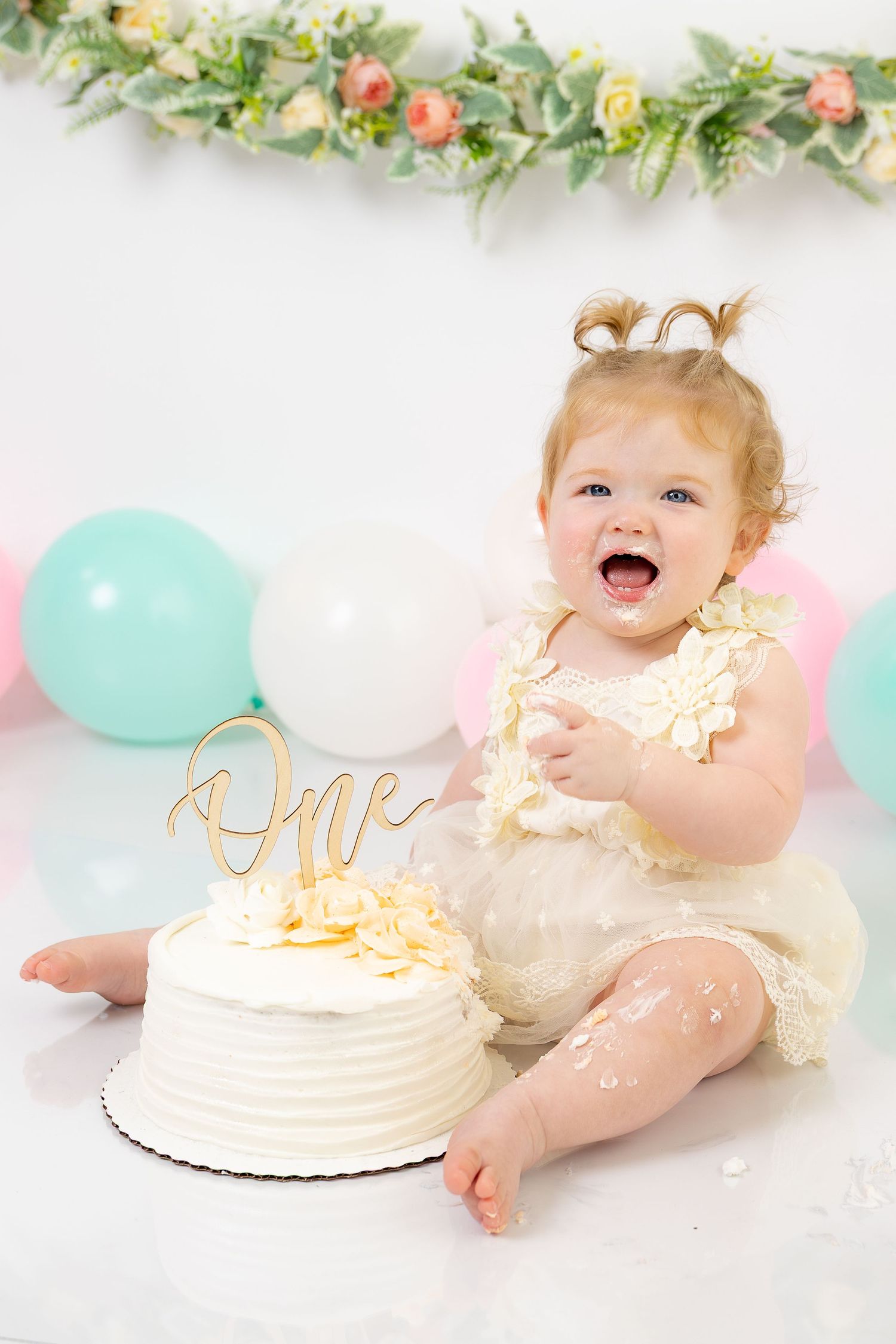 A joyful baby girl in a white dress celebrates her first birthday with a smash cake, balloons, and floral decor.