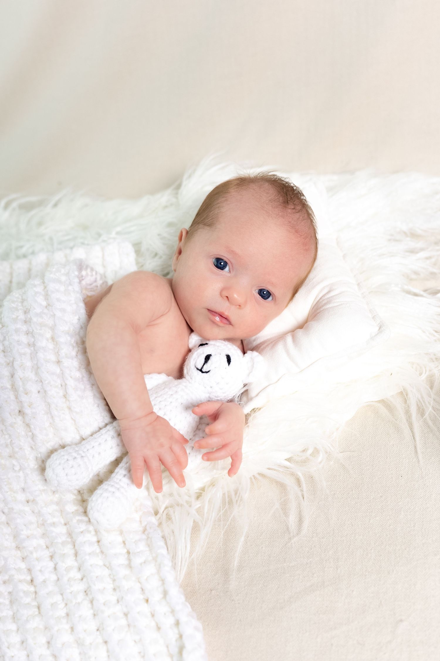 Baby lying on white blanket holding small panda toy with bright blue eyes.