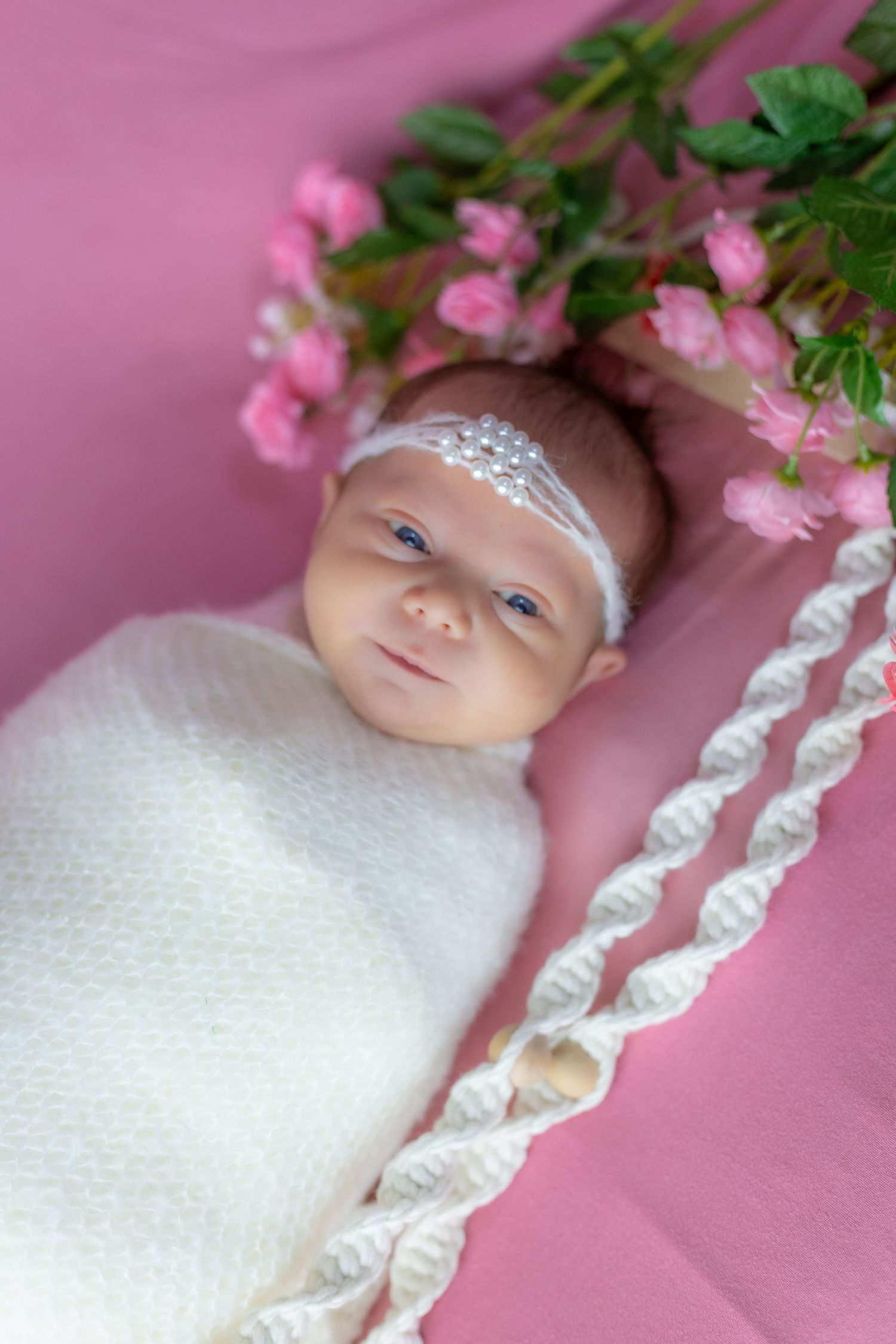 Baby in white swaddle and crystal headband lying on pink backdrop with delicate floral accents.