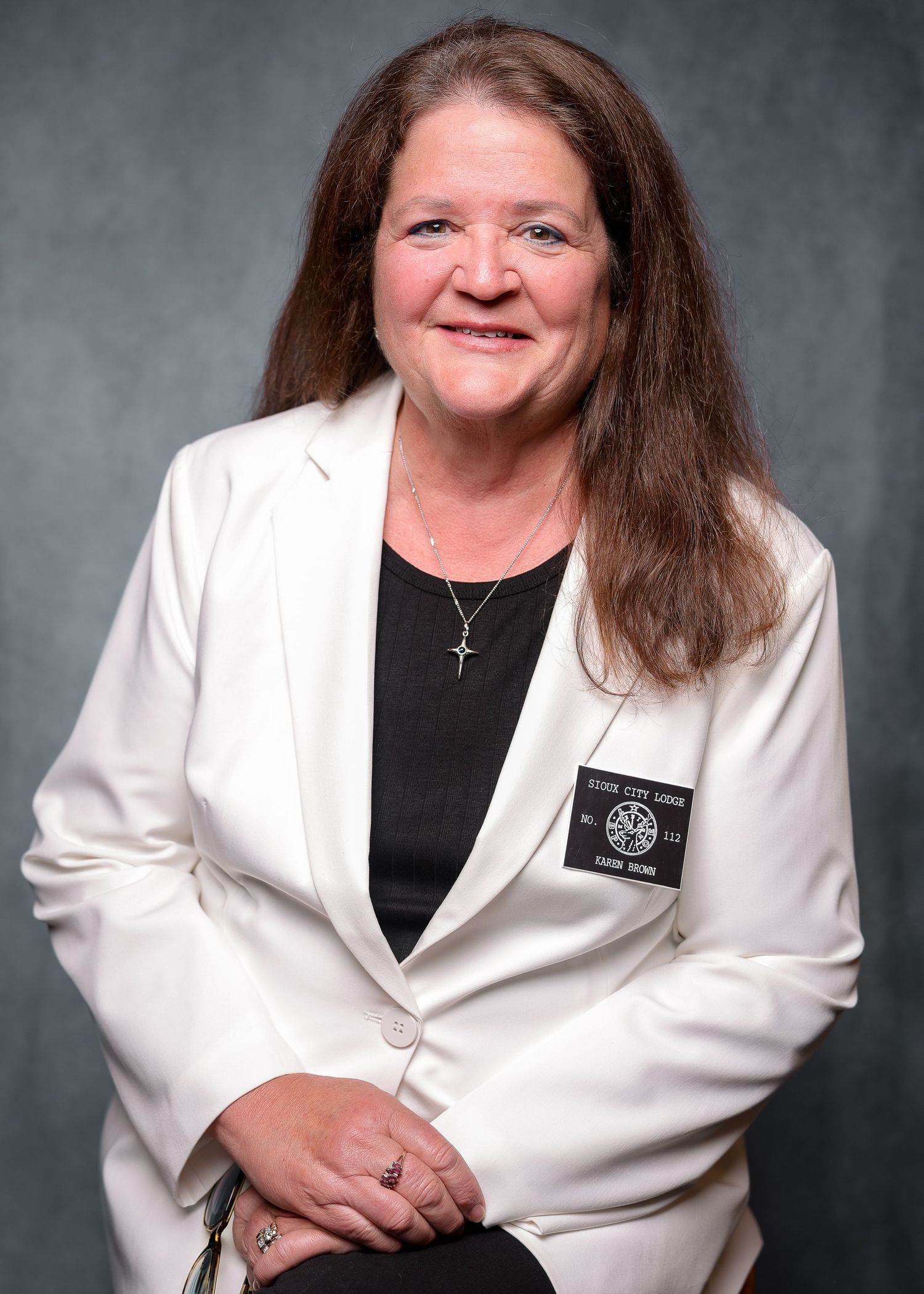 Woman in white blazer and black top with cross necklace and name badge smiling for professional headshot.
