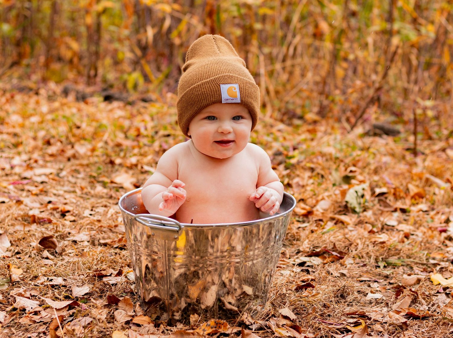 Baby wearing brown beanie sits in metal bucket surrounded by autumn leaves.