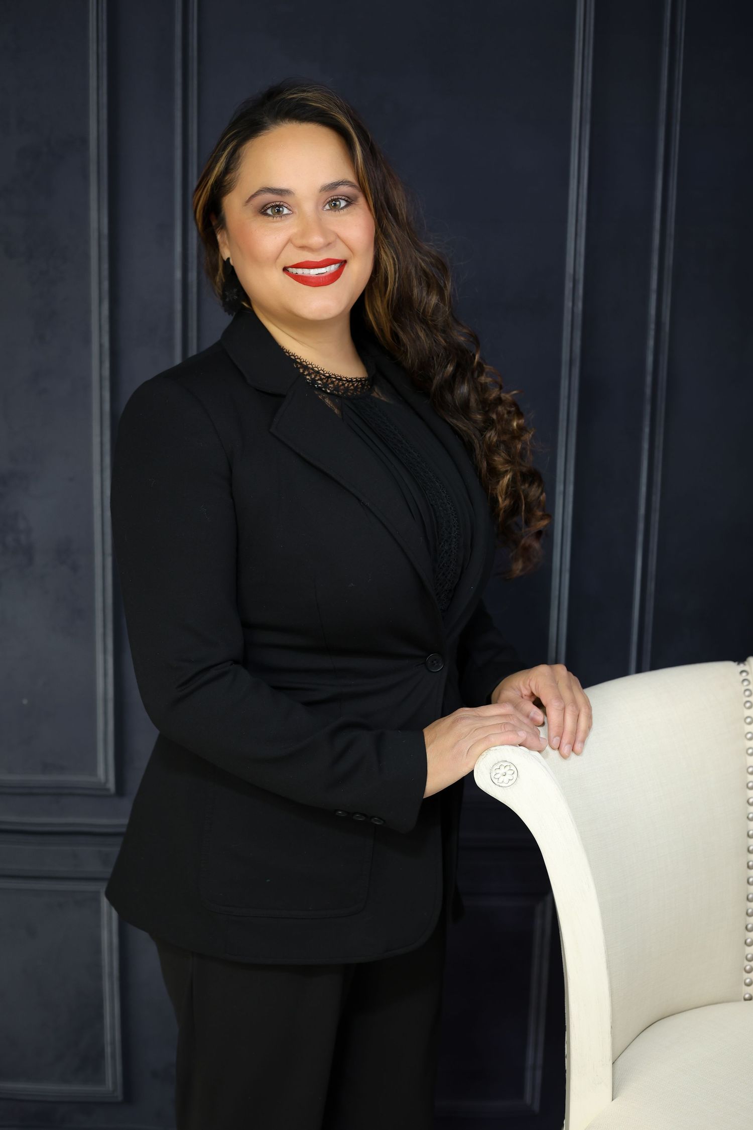 Professional portrait session with black attire and white chair against dark paneled wall.