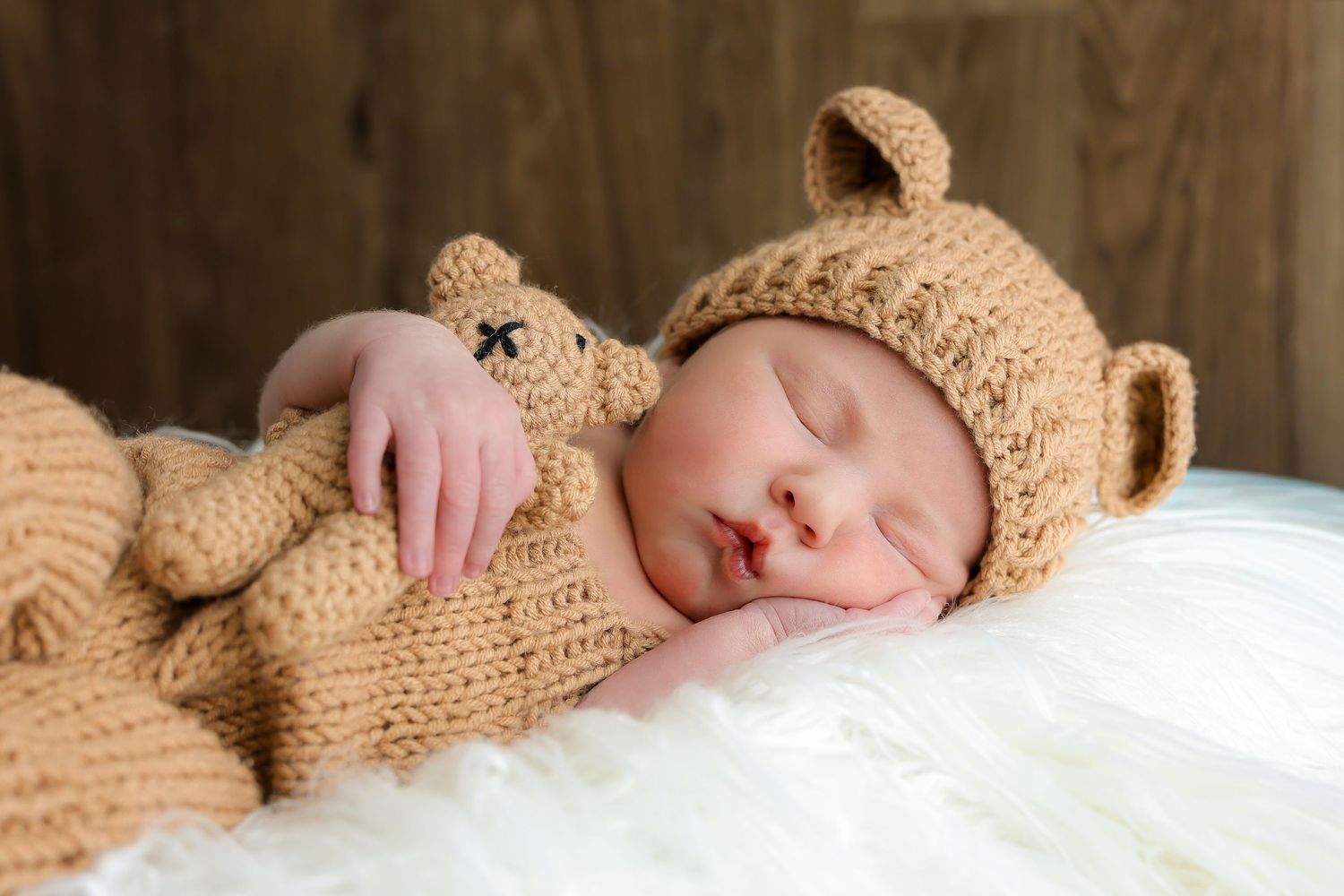 A sleeping newborn wearing a brown knit bear hat rests peacefully on white bedding.