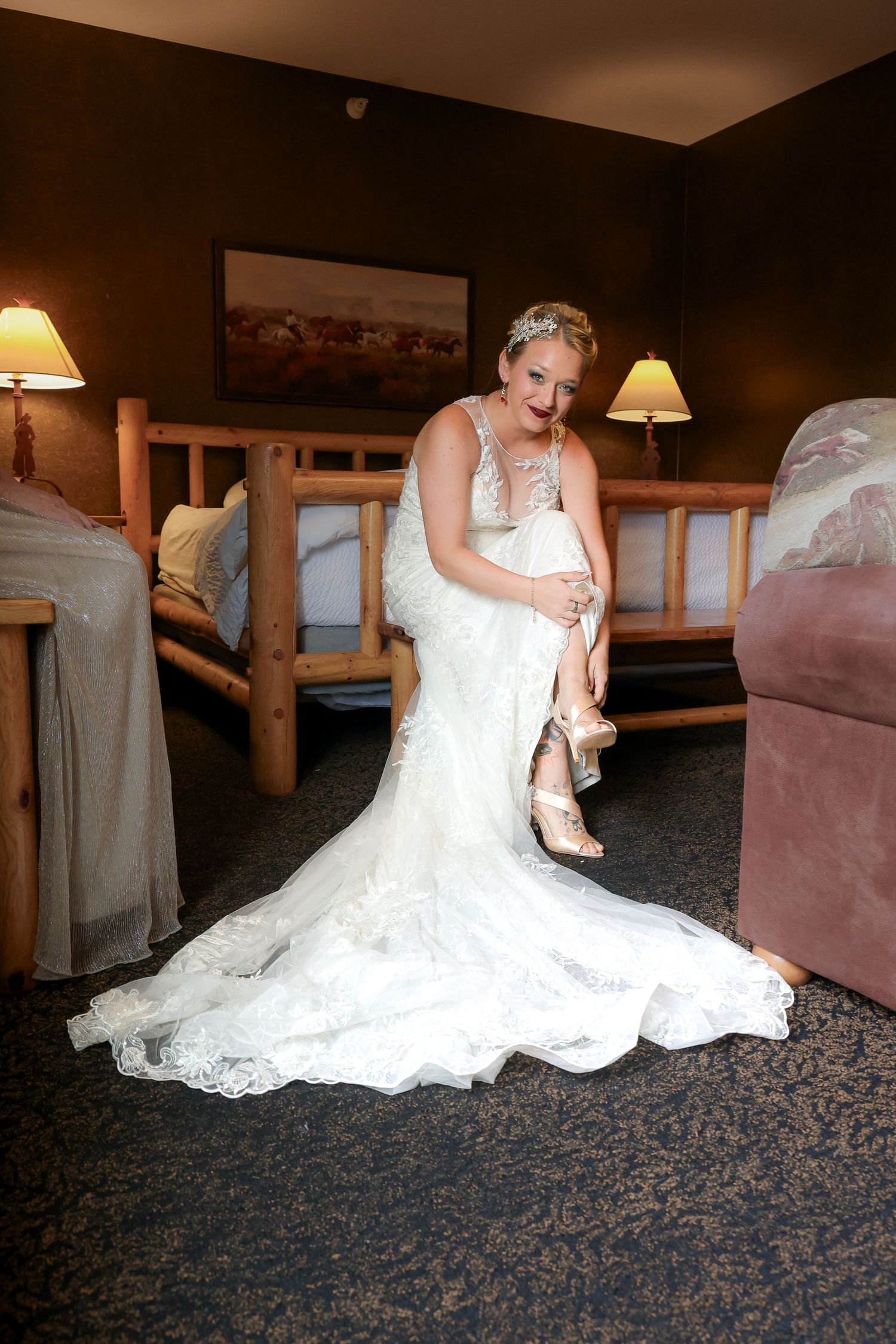 A bride in a white wedding gown adjusts her shoes while sitting on a patterned carpet in a dimly lit room.