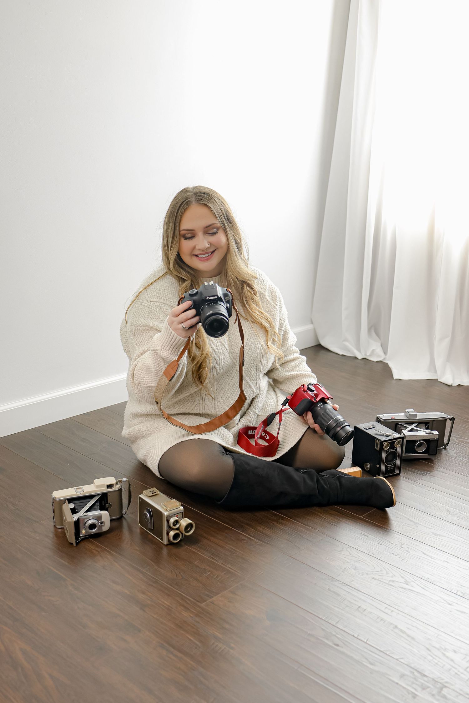 Smiling blonde woman sitting cross-legged on hardwood floor holding DSLR camera, surrounded by vintage cameras, wearing cream sweater.