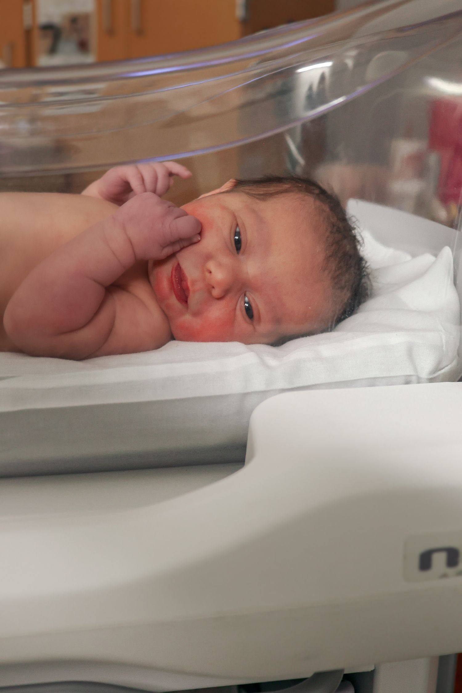 Newborn baby resting in clear hospital bassinet under warming lights.