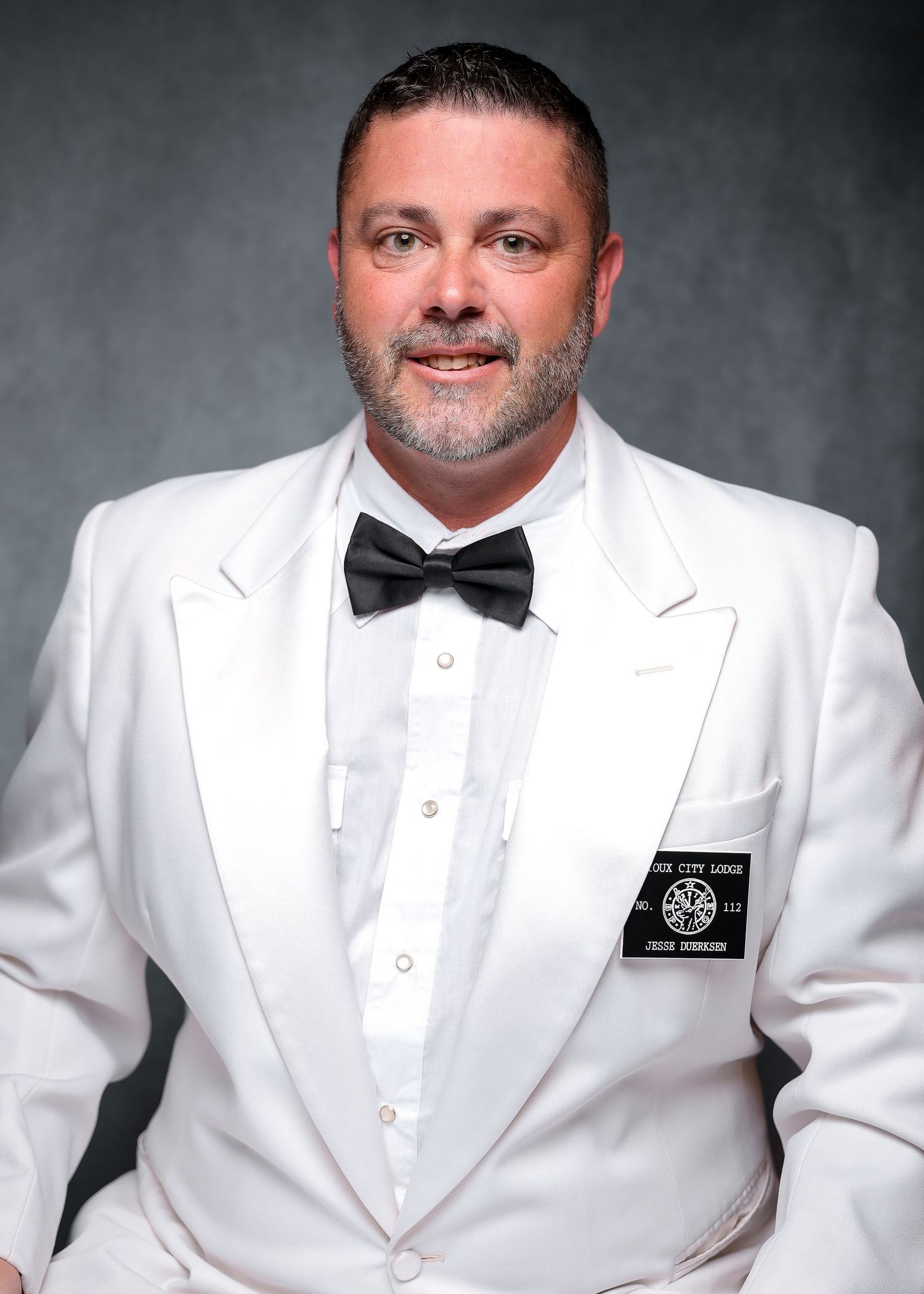 Man in white tuxedo jacket and black bow tie with a badge, posing for a formal portrait photo.