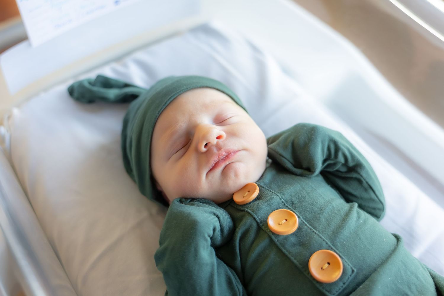 A baby in a green button-up sleeper lies on white bedding with their eyes closed in a peaceful sleeping position.