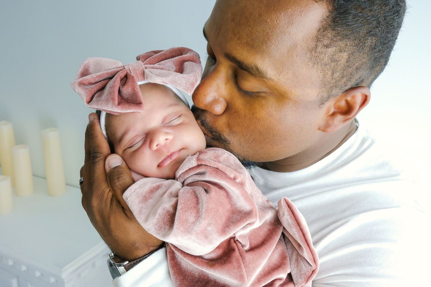A parent in a white shirt tenderly kisses their newborn baby wrapped in a pink swaddle blanket.