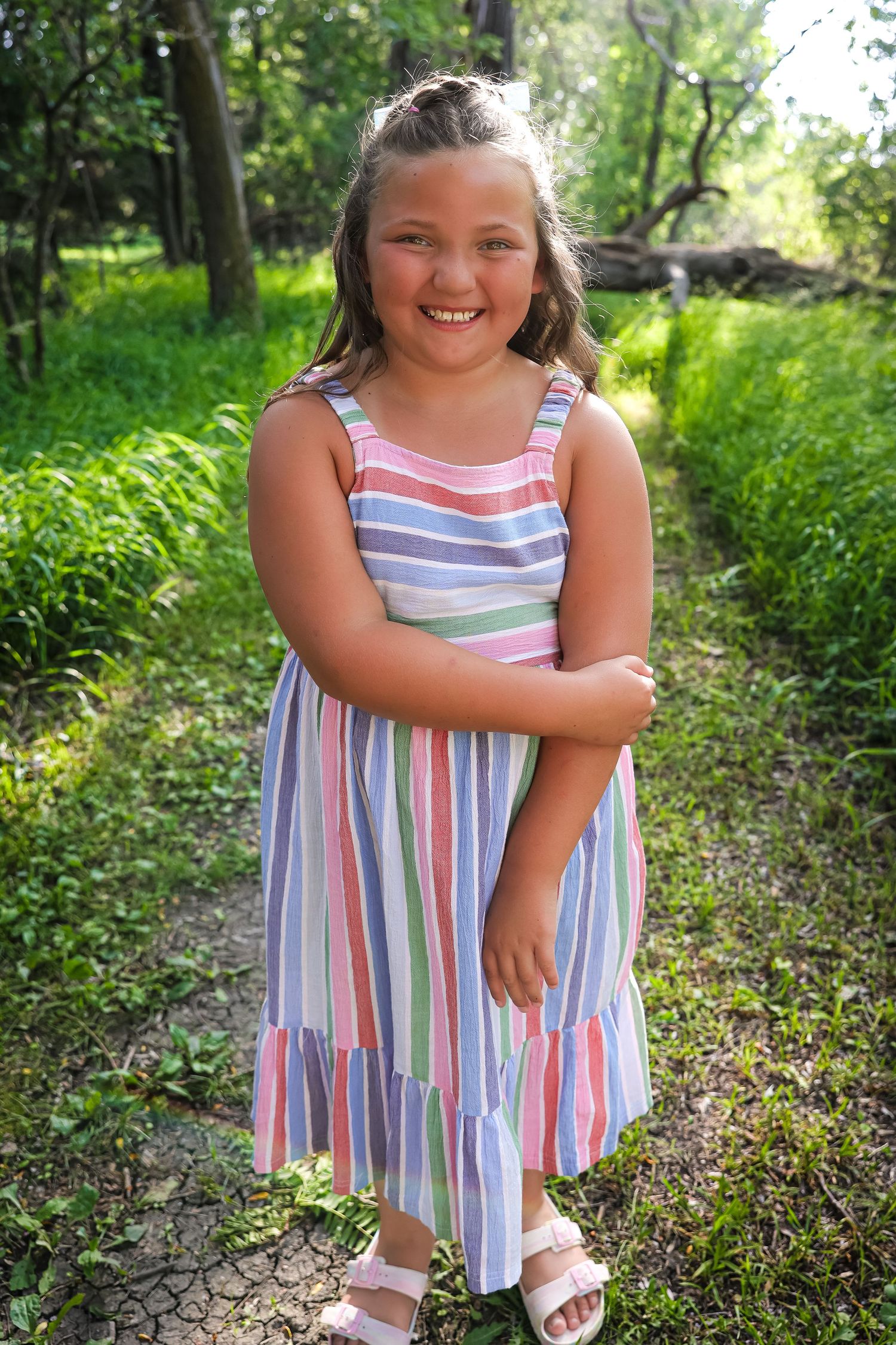 Young person in striped summer dress poses playfully in outdoor woodland setting.