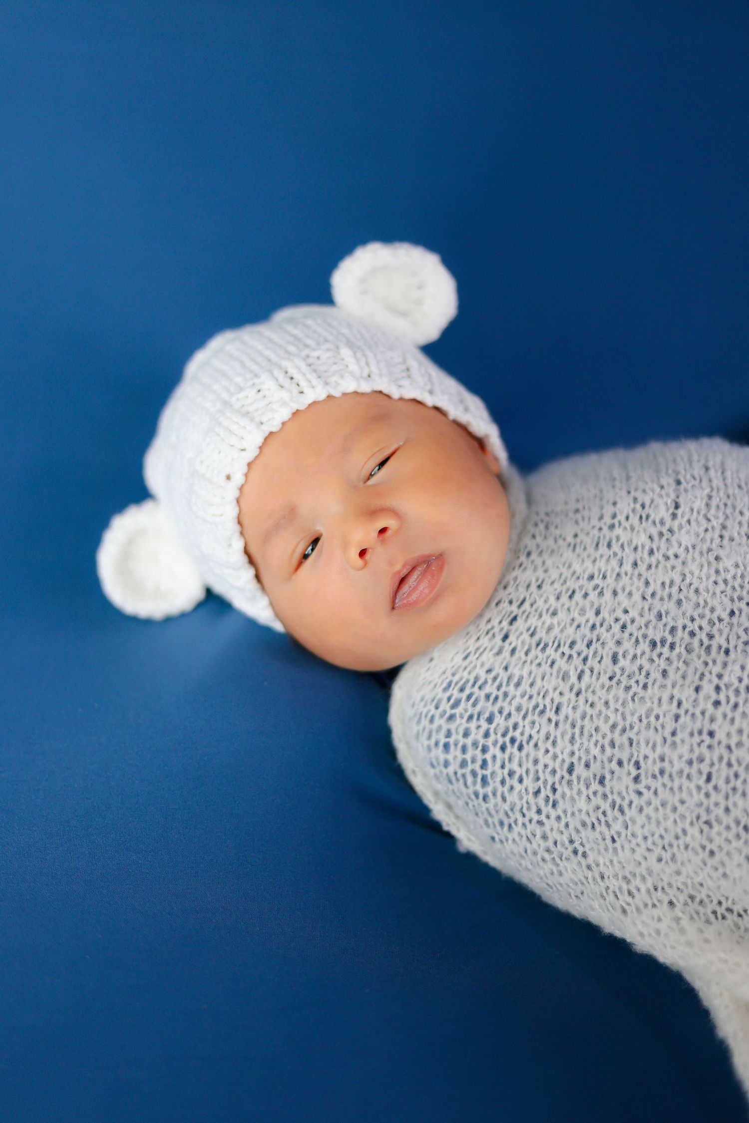 Peaceful newborn wearing white knit hat against deep blue background.