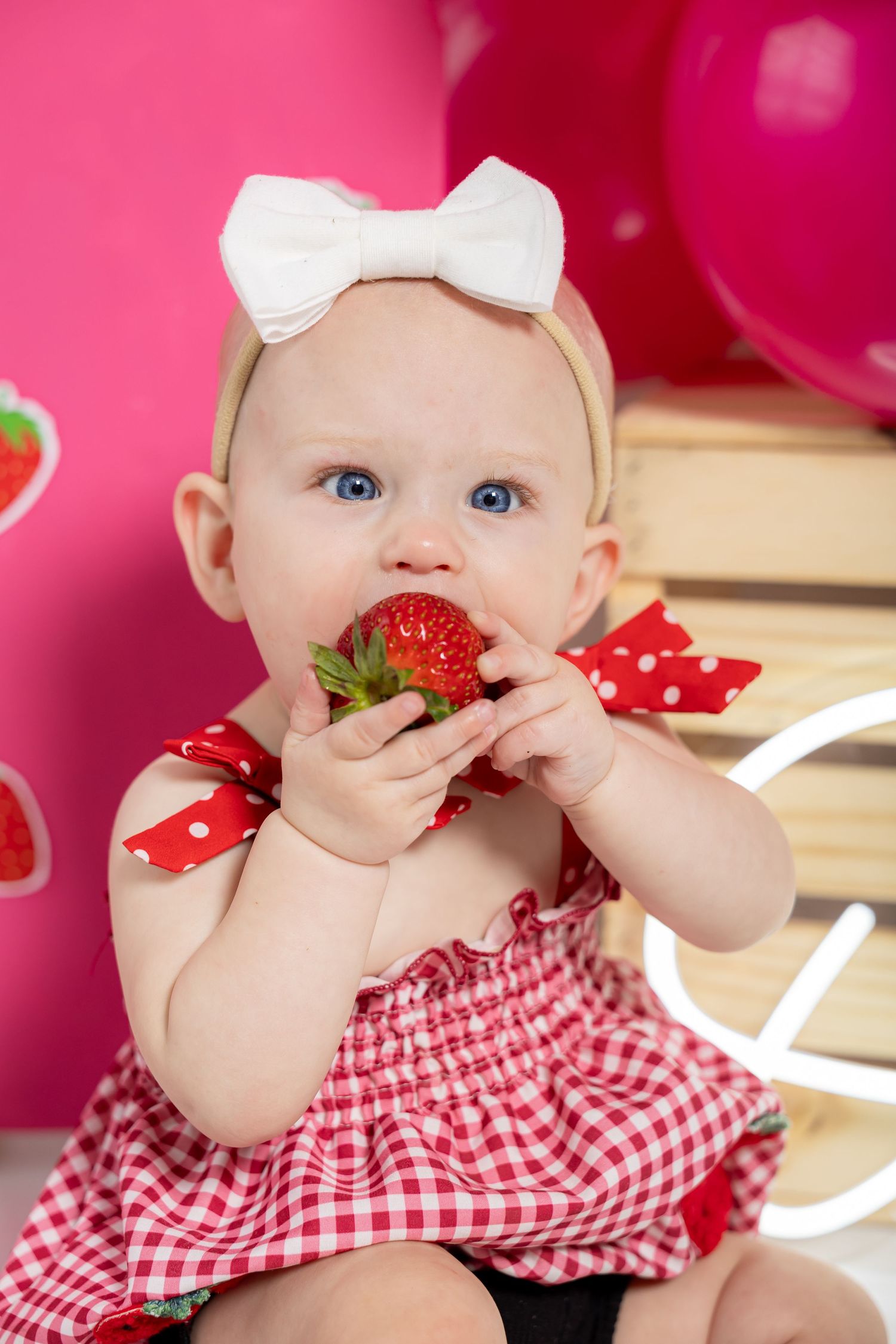 Red gingham outfit and strawberry themed cake smash photo against pink background.