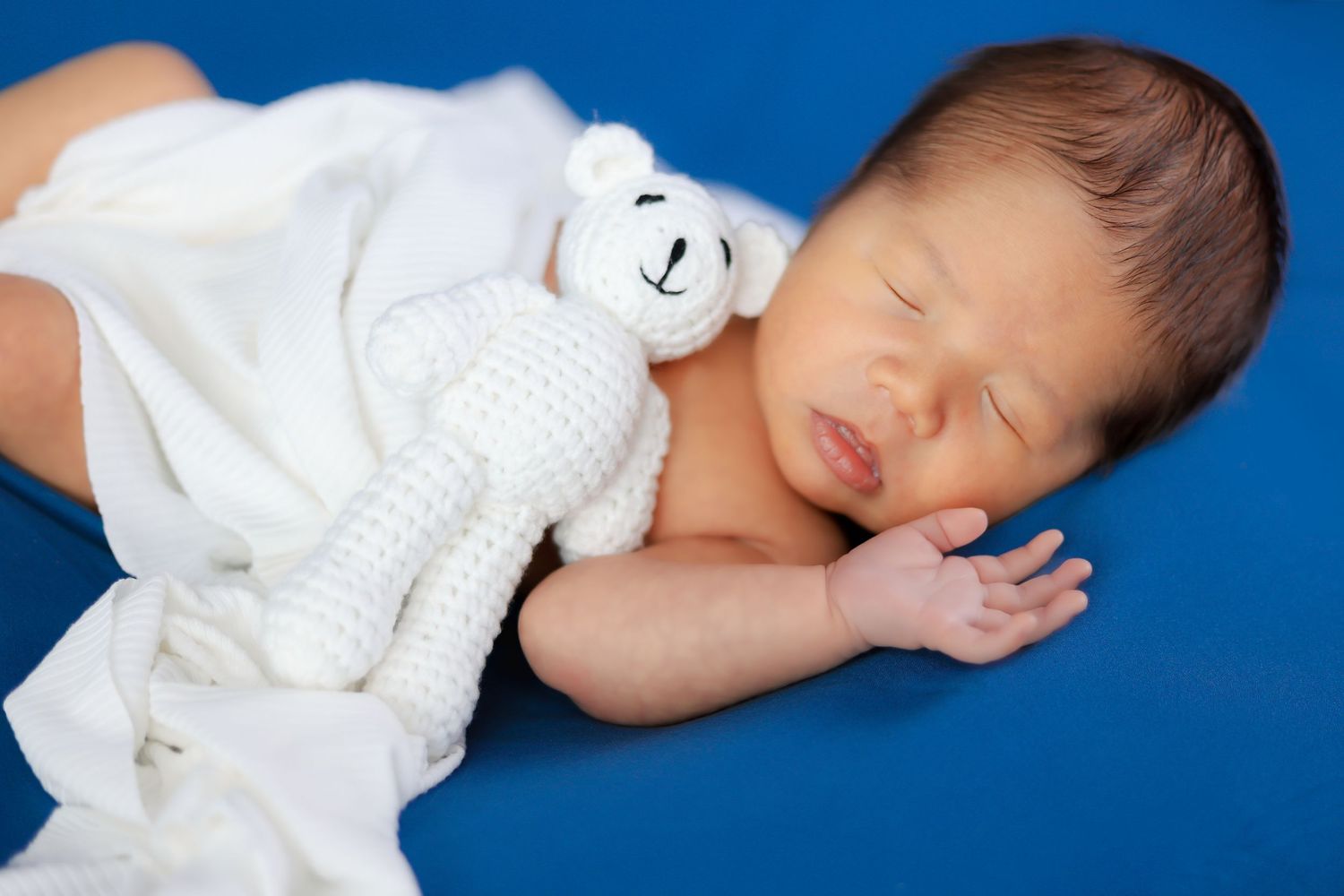 Newborn sleeping peacefully with white teddy bear against blue background.