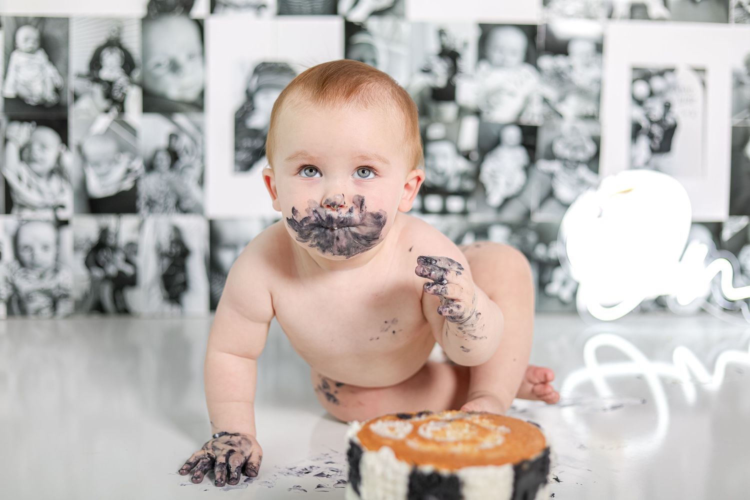 A baby with frosting on their face reaches for a cake during a smash cake session.