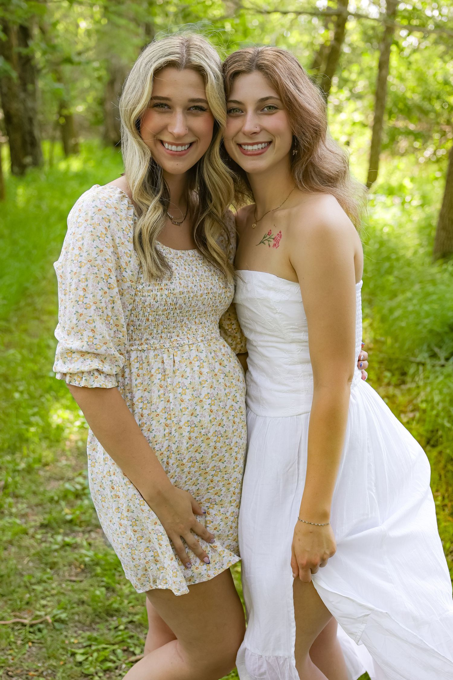 Two friends in summer dresses pose together in a sunny forest setting with natural lighting and greenery.
