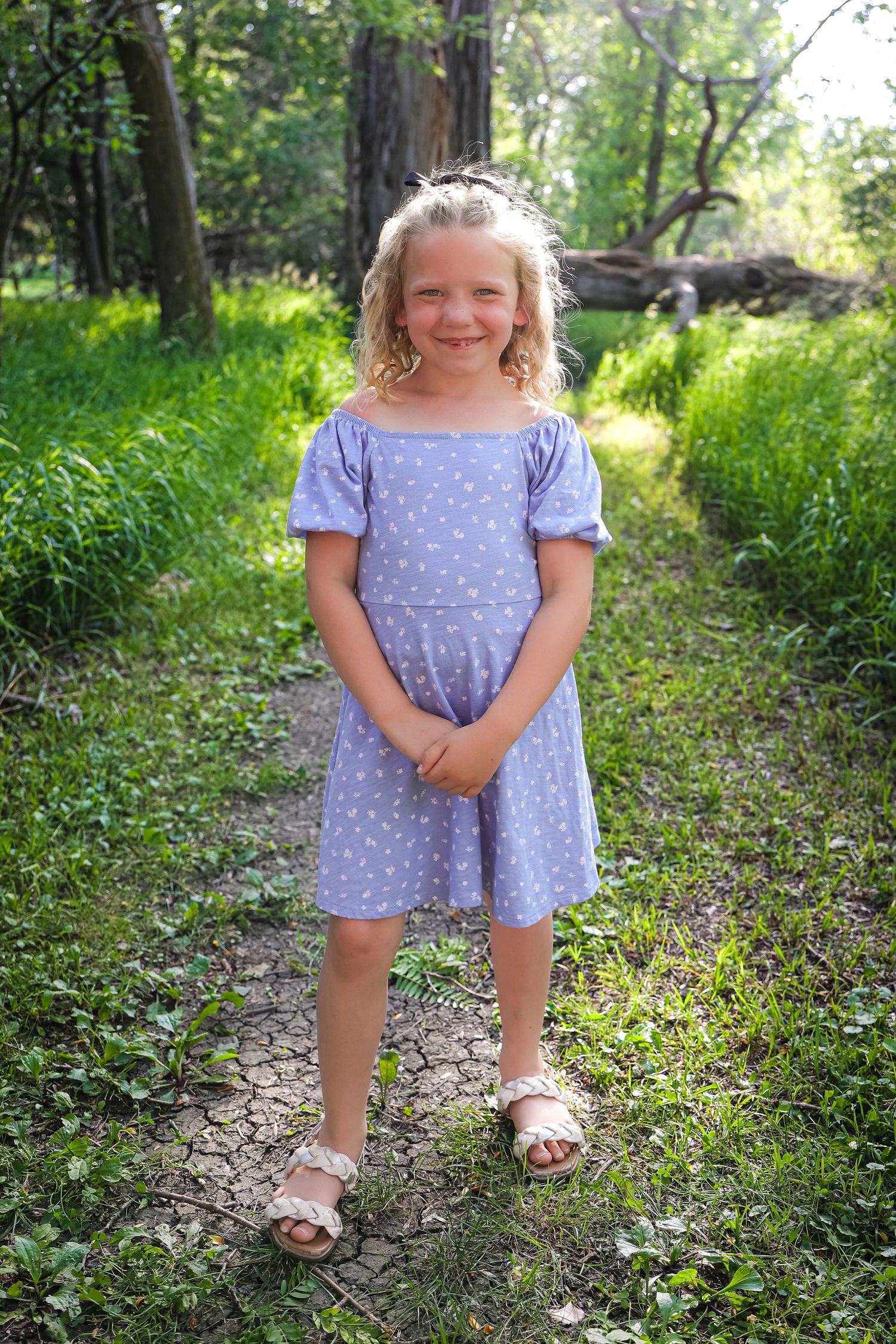 Child in purple polka dot dress poses playfully on woodland path.