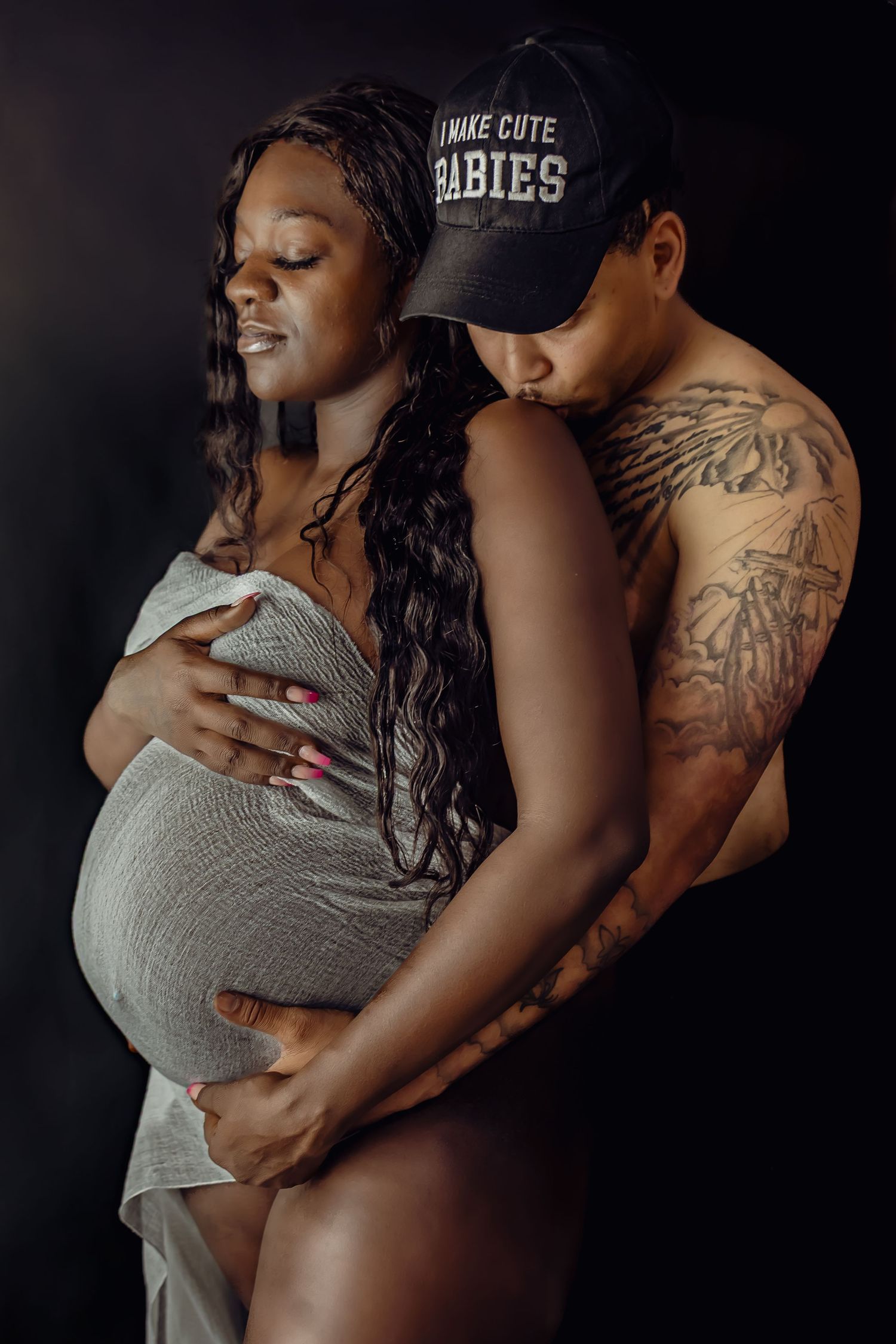 Intimate maternity portrait of expecting parents embracing against dark background.