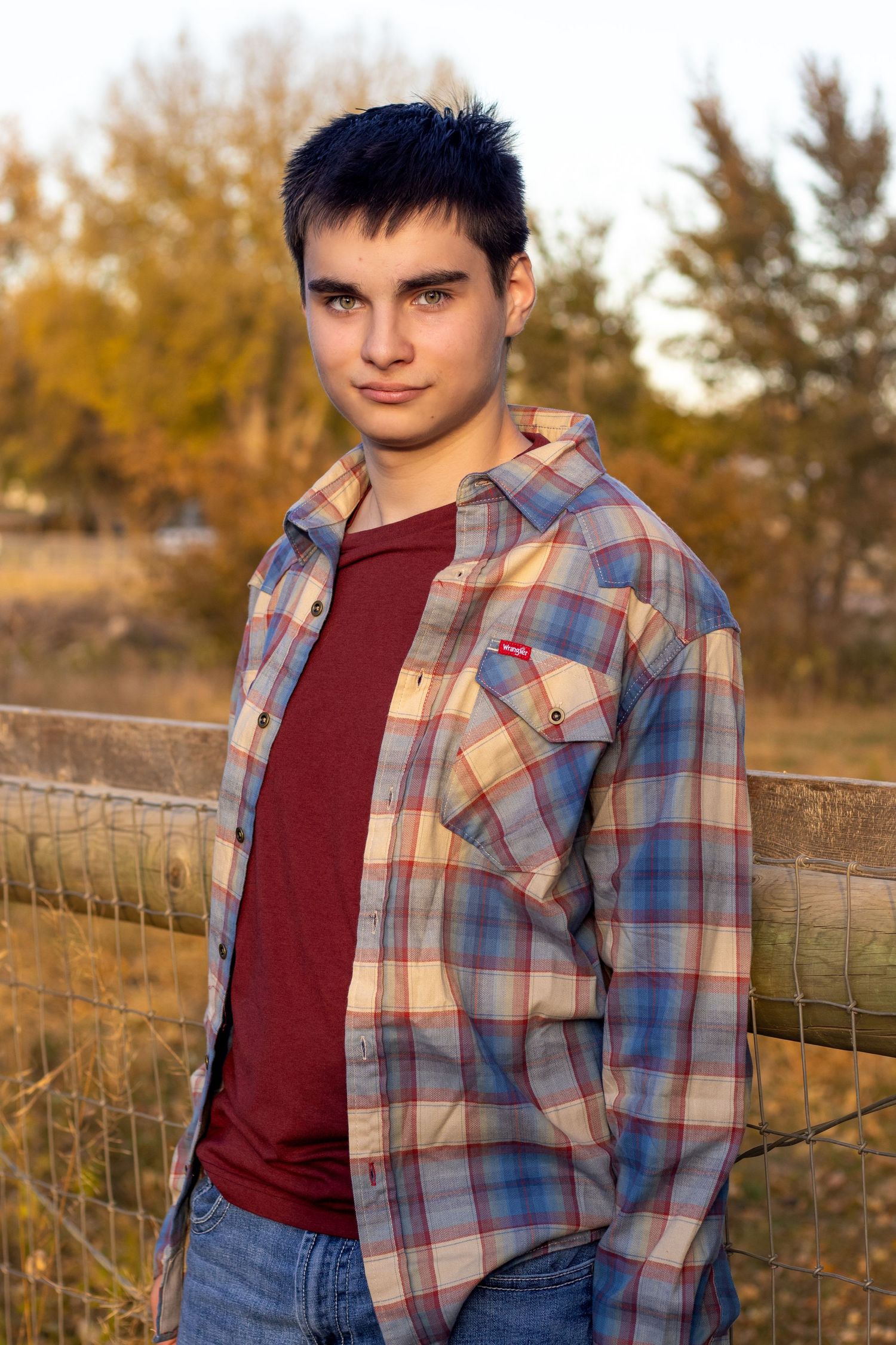A person in a plaid shirt and burgundy tee poses against an autumn landscape.