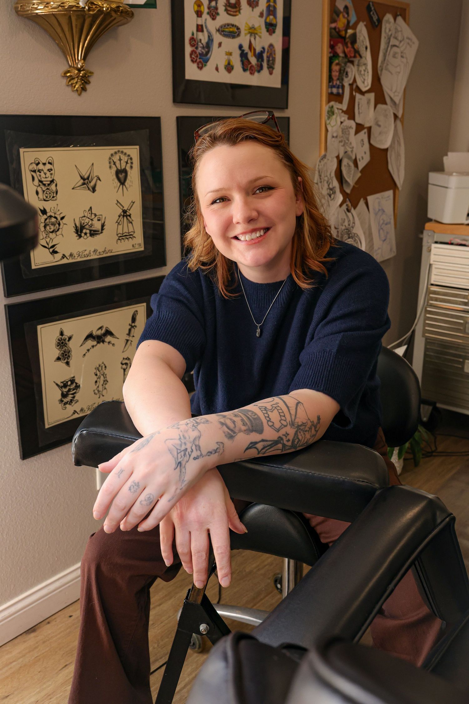 Smiling tattoo artist sits in studio chair showing forearm tattoos, with framed flash art on walls behind her.