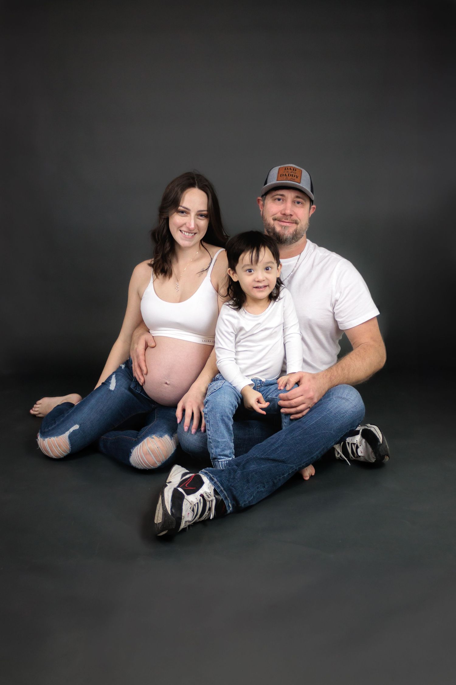 Family maternity portraits with casual attire sitting on dark floor.