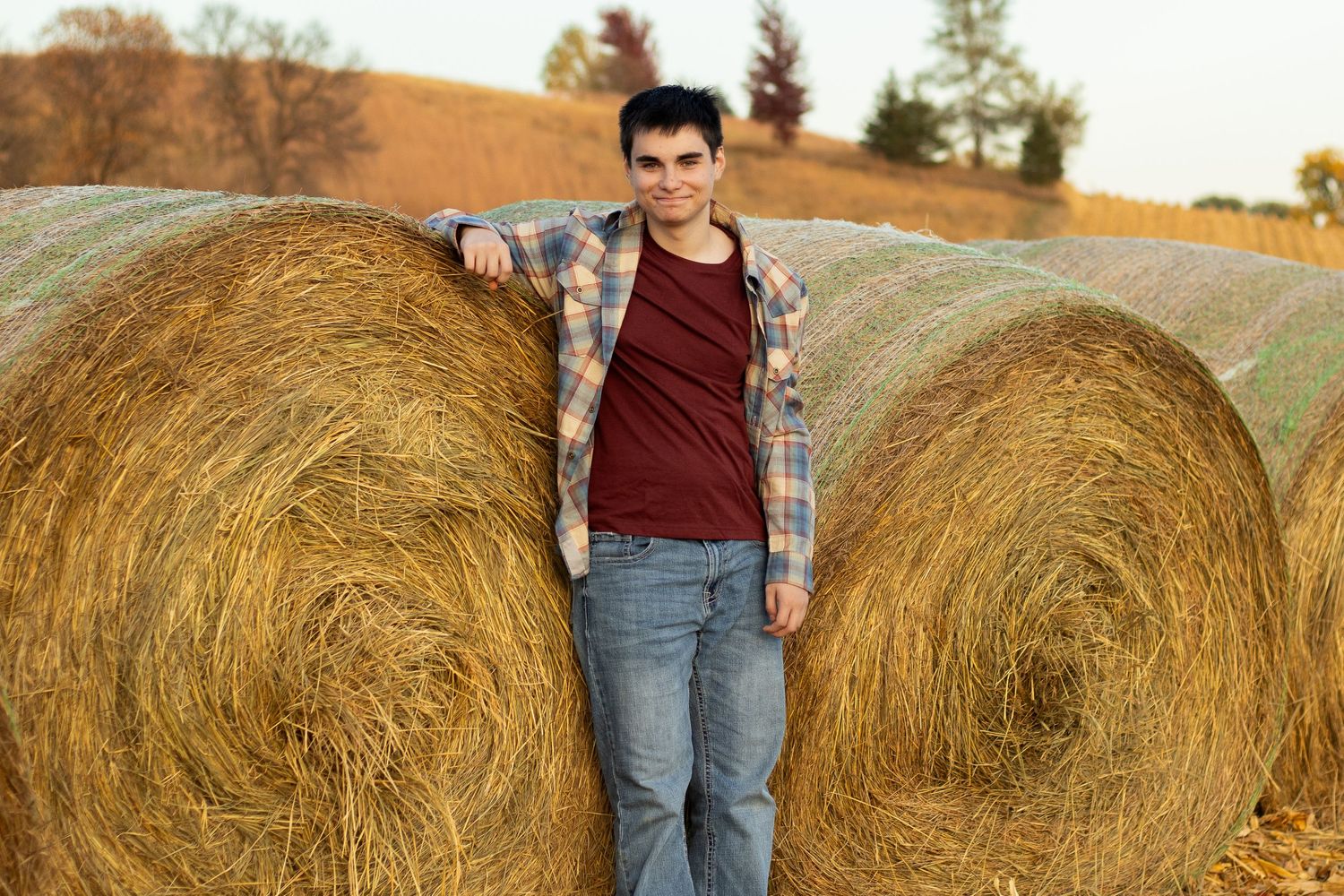 A person in plaid and jeans poses against round hay bales in a rural autumn setting.