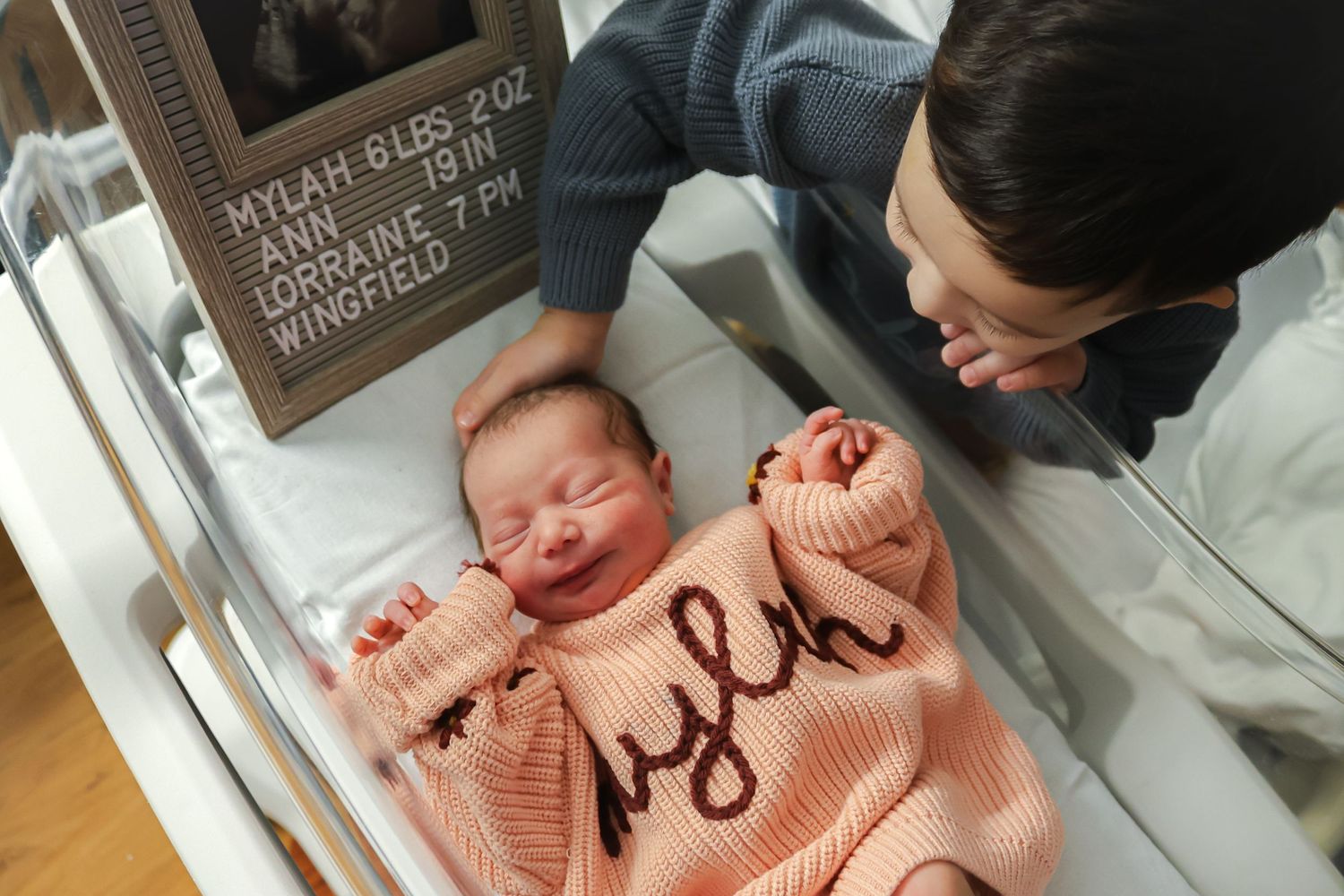 Newborn baby wearing a peach-colored sweater in a hospital bassinet.
