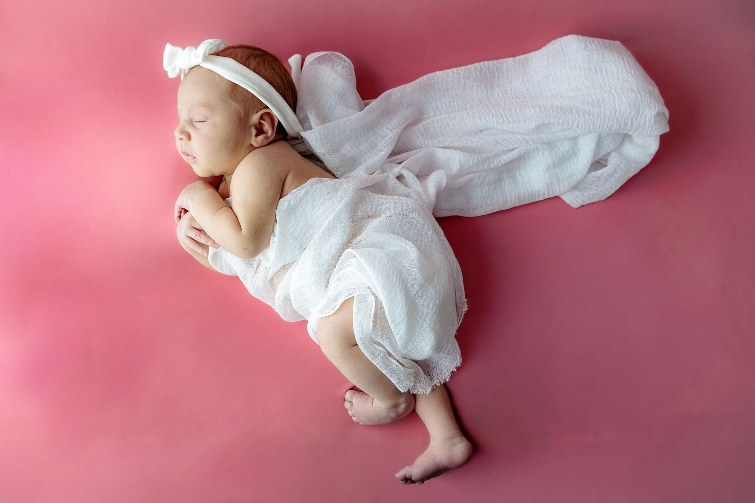 A sleeping baby wearing a white onesie lies on a pink background in a peaceful pose.