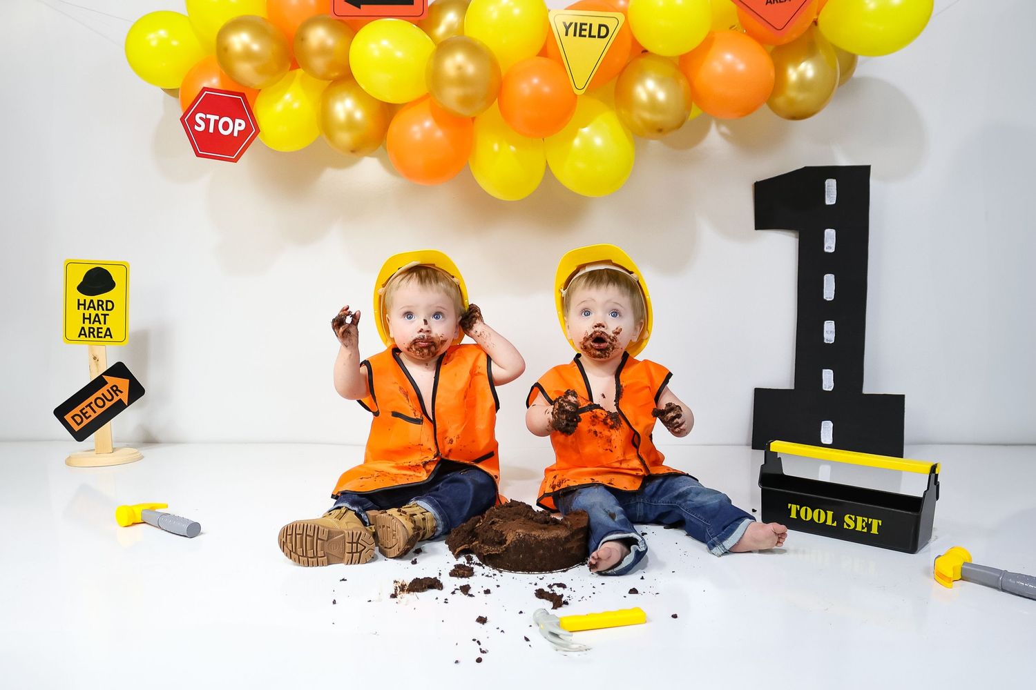 Construction themed photo setup with orange and yellow balloons and props against white backdrop.