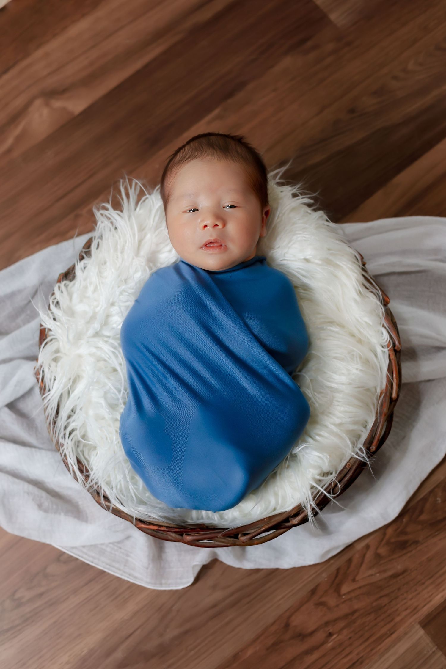 Newborn wrapped in blue swaddle resting in white nest on wooden floor.