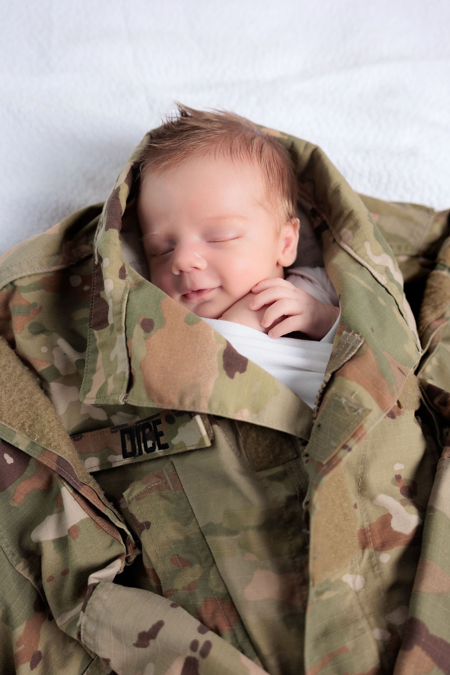 A sleeping baby wrapped in camouflage military fatigues with name tape visible.