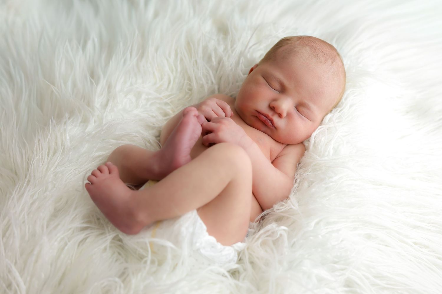 Newborn baby sleeping peacefully on soft white fur blanket in natural light.